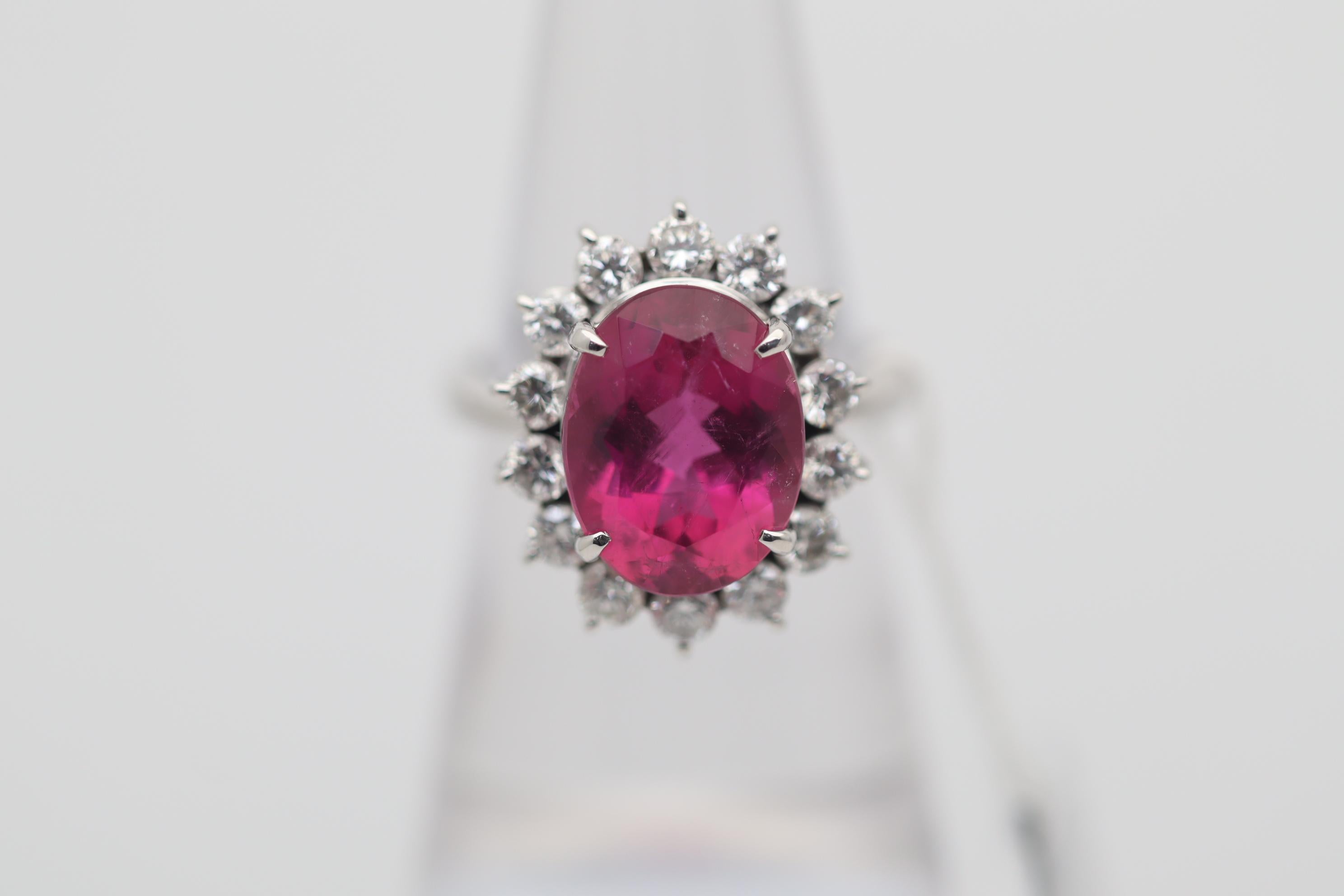 A sweet and stylish platinum ring featuring a bright rich pink tourmaline weighing 5.00 carats. It has a lovely oval-shape and a reddish-pink color that some consider to be rubellite. It is complemented by a halo of round brilliant-cut diamonds