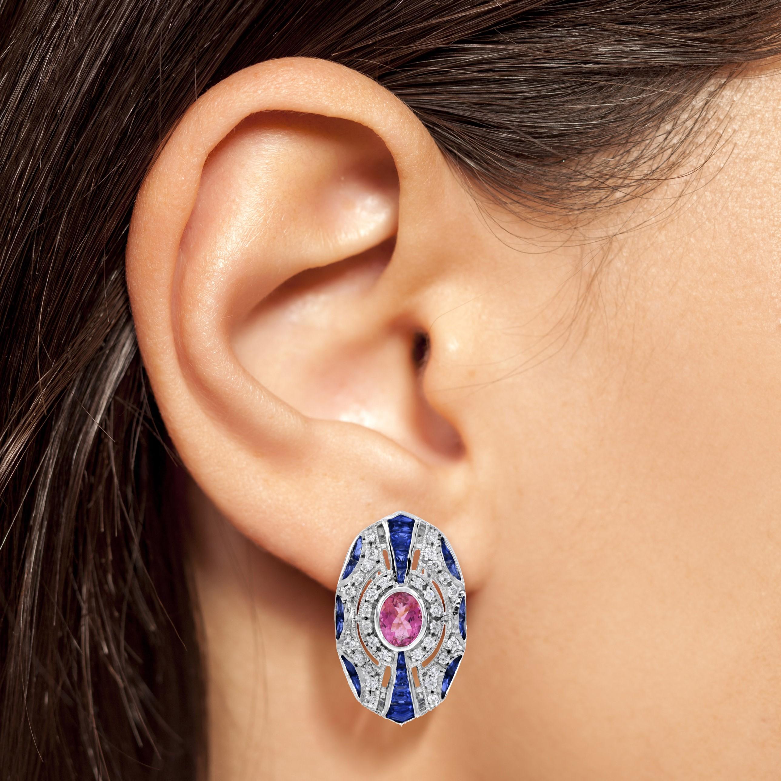 A pink tourmaline, weighing approx. 1.10 carats each side, glistens and glows in the center of geometrically constructed Art Deco style omega back earrings. The gemstone is surrounded by round diamonds and French cut sapphires accented with