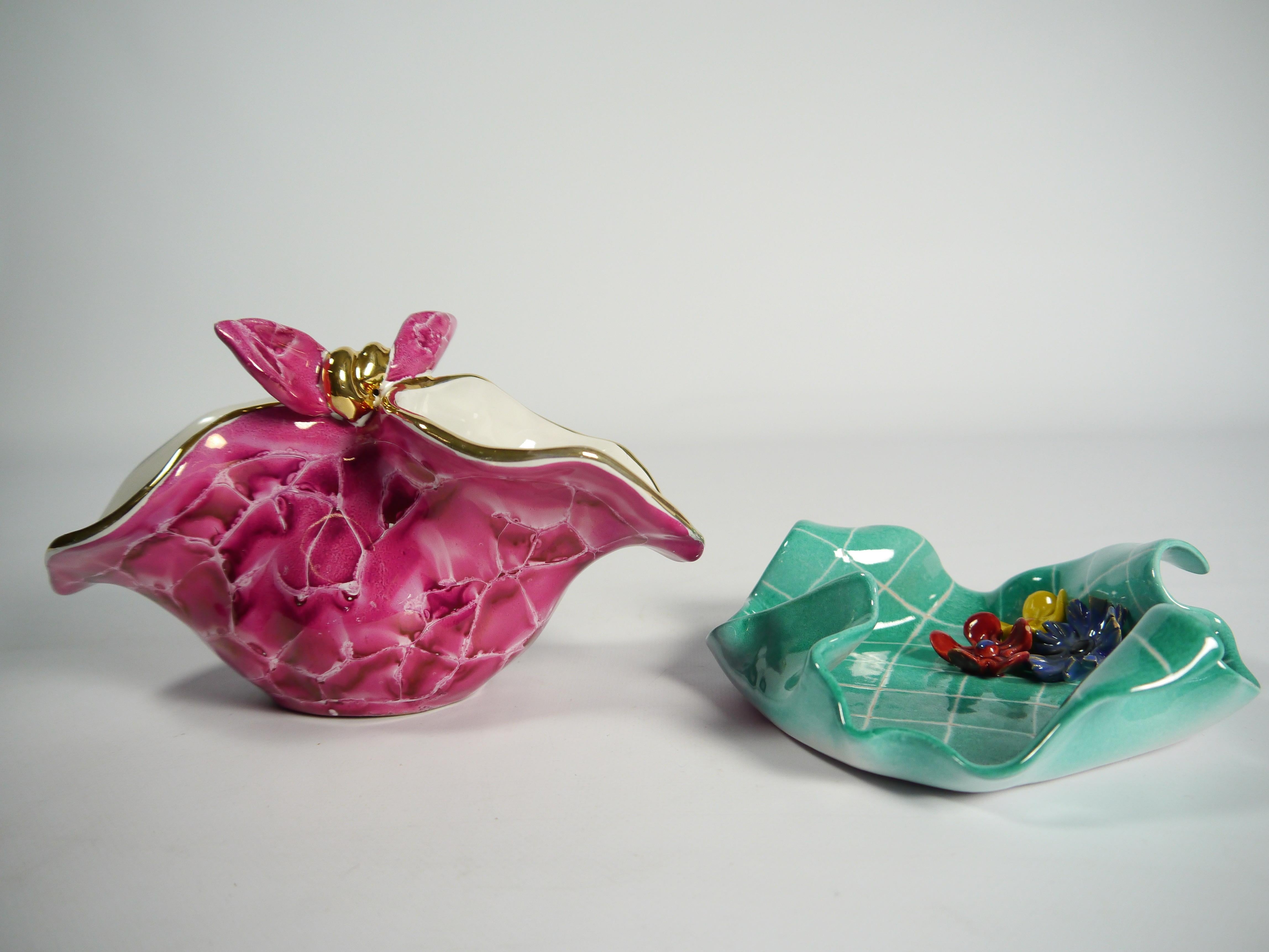 Two Napkin-shaped ceramic dishes; one pink/golden bowl/dish imitating a tied napkin, one turquoise platter ornamented with flowers imitating a napkin with raised corners. Kitschy in its appearances and colors.  

Size refers to the pink/golden dish.