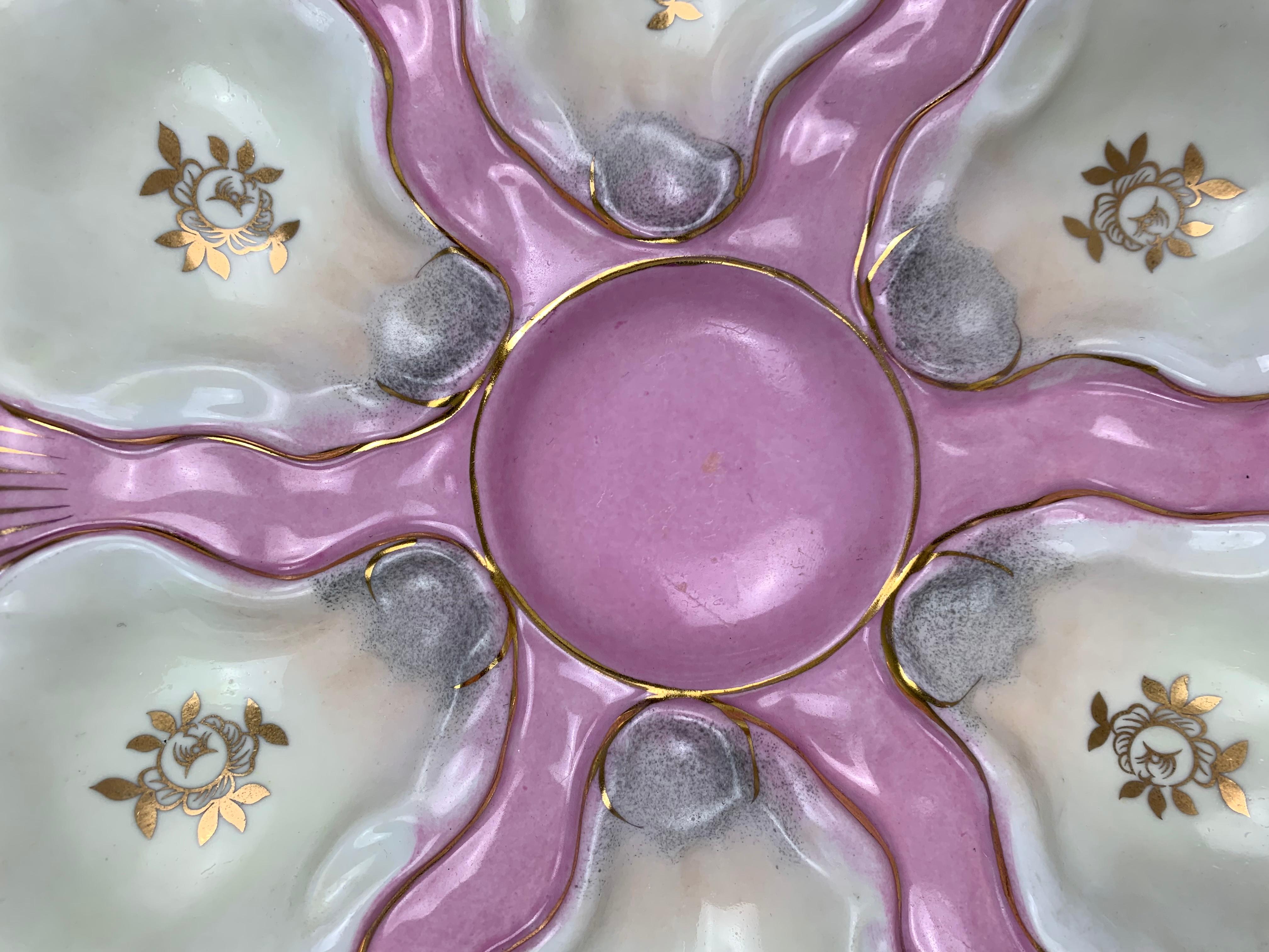 Gorgeous porcelain pink and white 20th Century oyster plates with six oyster wells bordered in gold gilt. Gold gilt flower in each of the wells.  Plates are footed with two holes for wall mounting. 