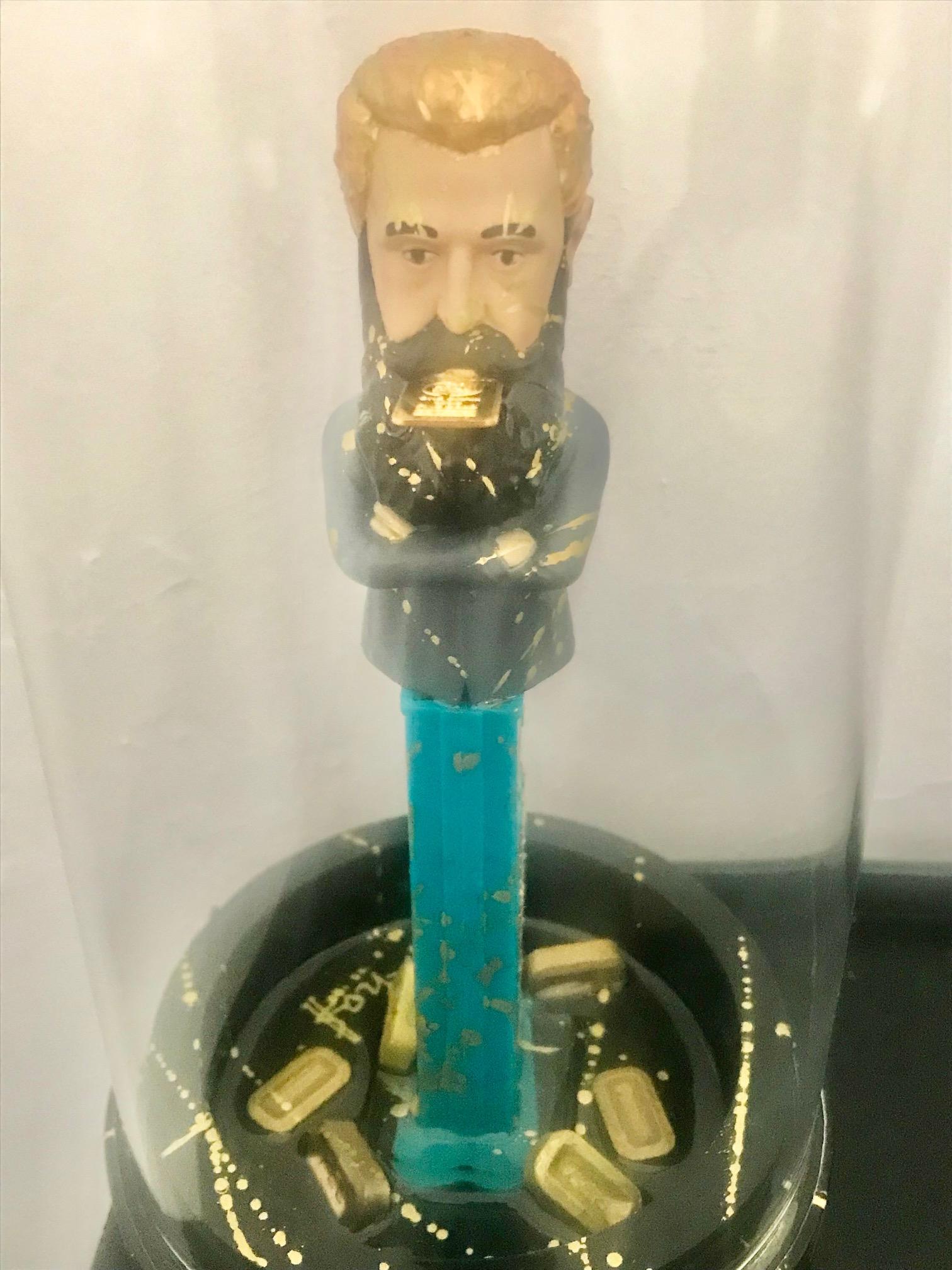 Sculpture combining a glass box displaying a plastic/resin character enhanced with acrylic paint and real customized candies are scattered around the base of the sculpture.

Instead of delivering candies the mouth of the character exhibits a real