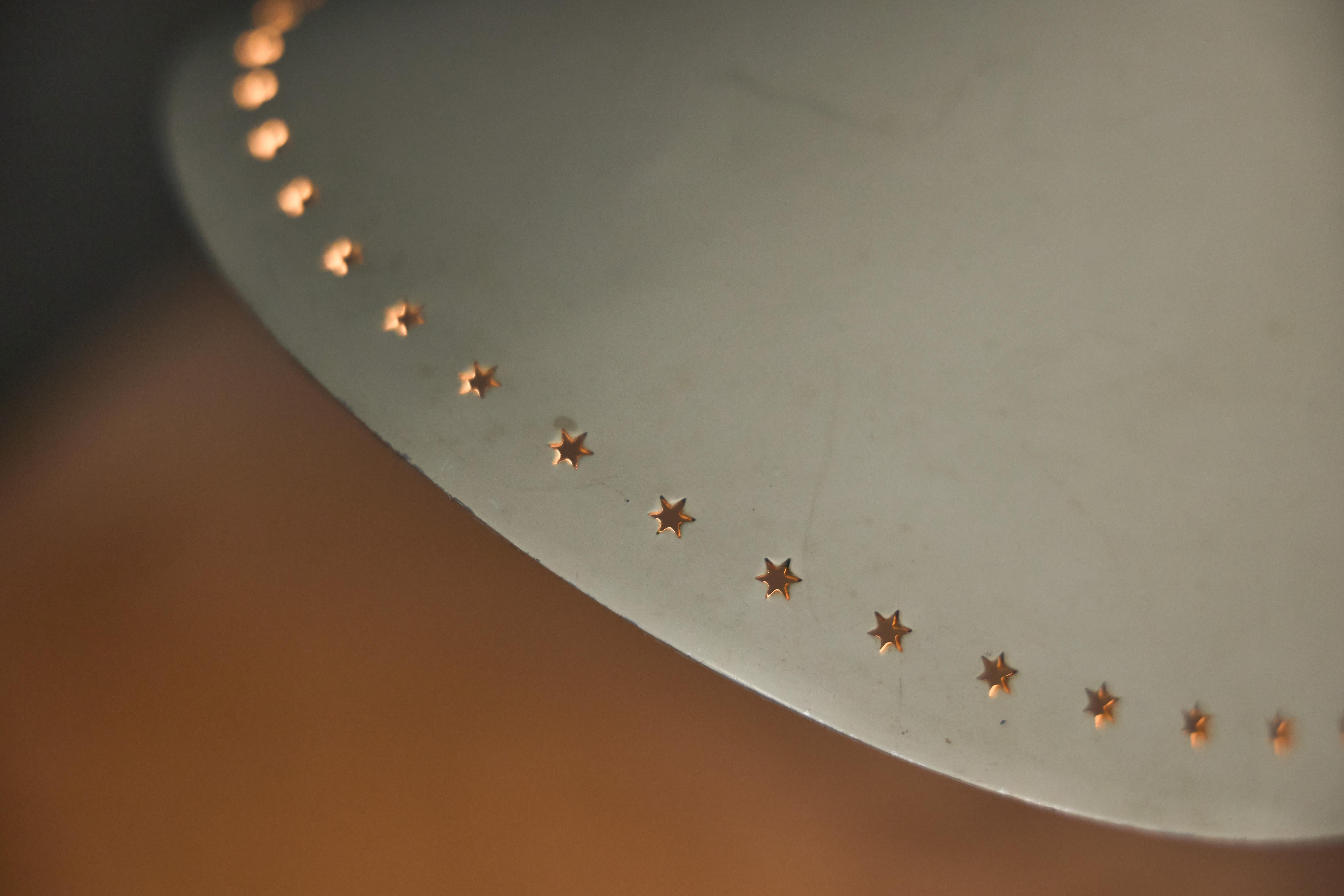 Designed by Herman Busquet for Hala Zeist in 1956, this Pinocchio table lamp features an adjustable metal frame with a sleek, off-white conical shade. The perforated star pattern along the edge creates a warm and atmospheric light effect when