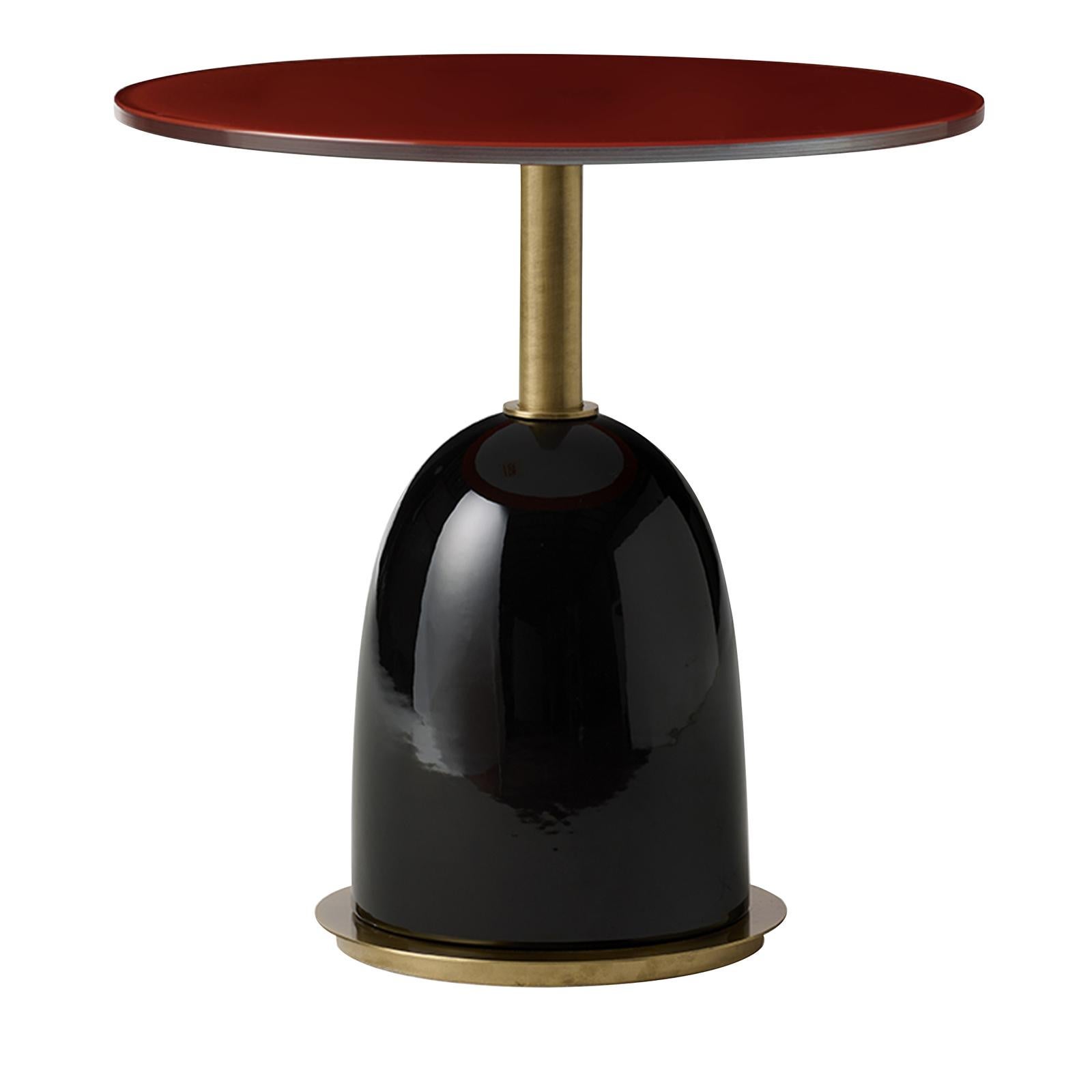 This side table features a structure in brass with a curved base covered in enamel-finished ceramic with a black color that strikingly contrasts with the vivid red of the round top crafted of crystal. The top is adorned with a series of square
