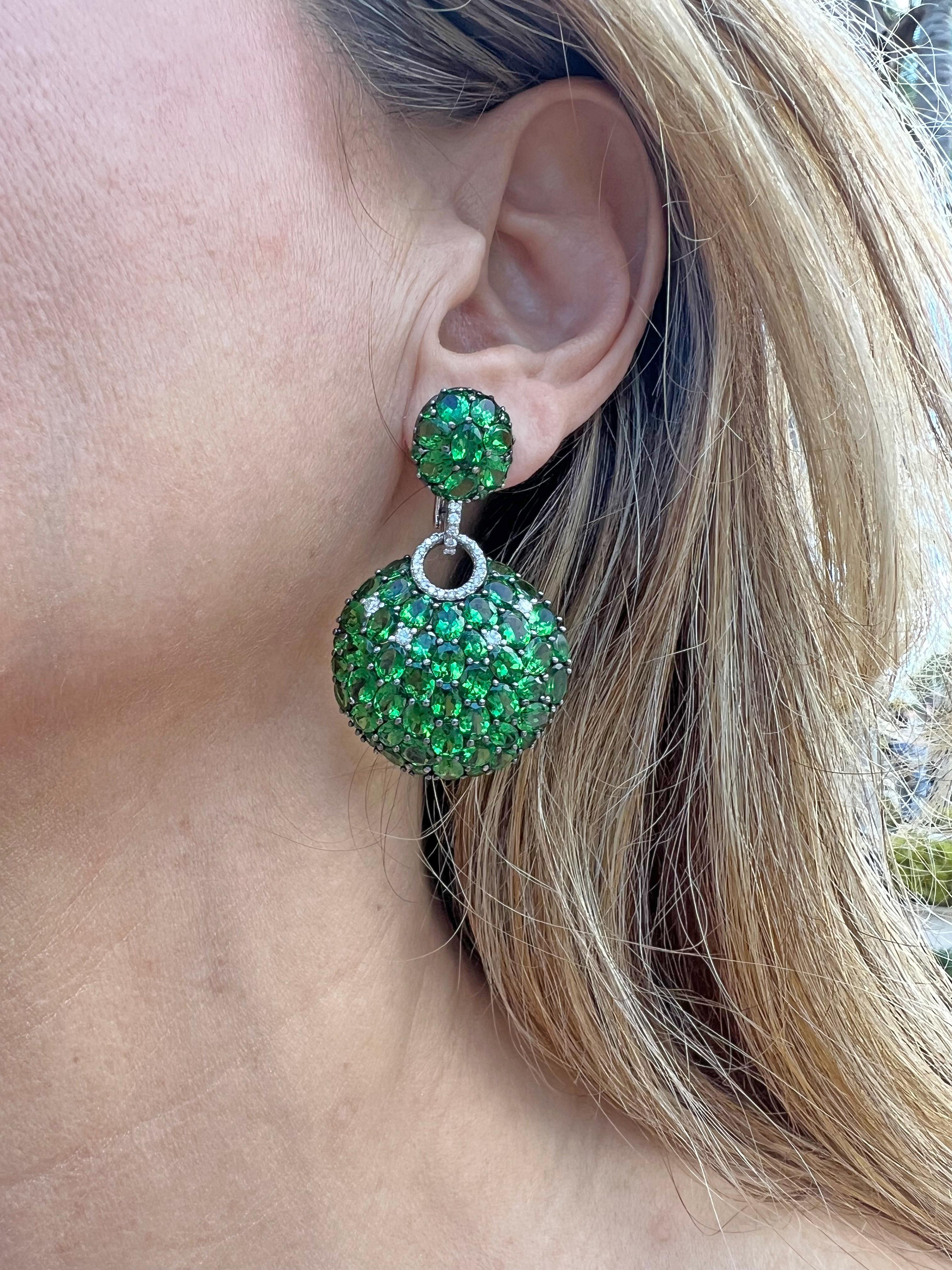 Tsavorite garnet and diamond pendant earrings in polished 18k white gold.  Larger circular-shaped domed drops suspended from smaller oval-shaped domed tops, both pave-set with tsavorite garnets and diamonds throughout.  Joined in the center by a
