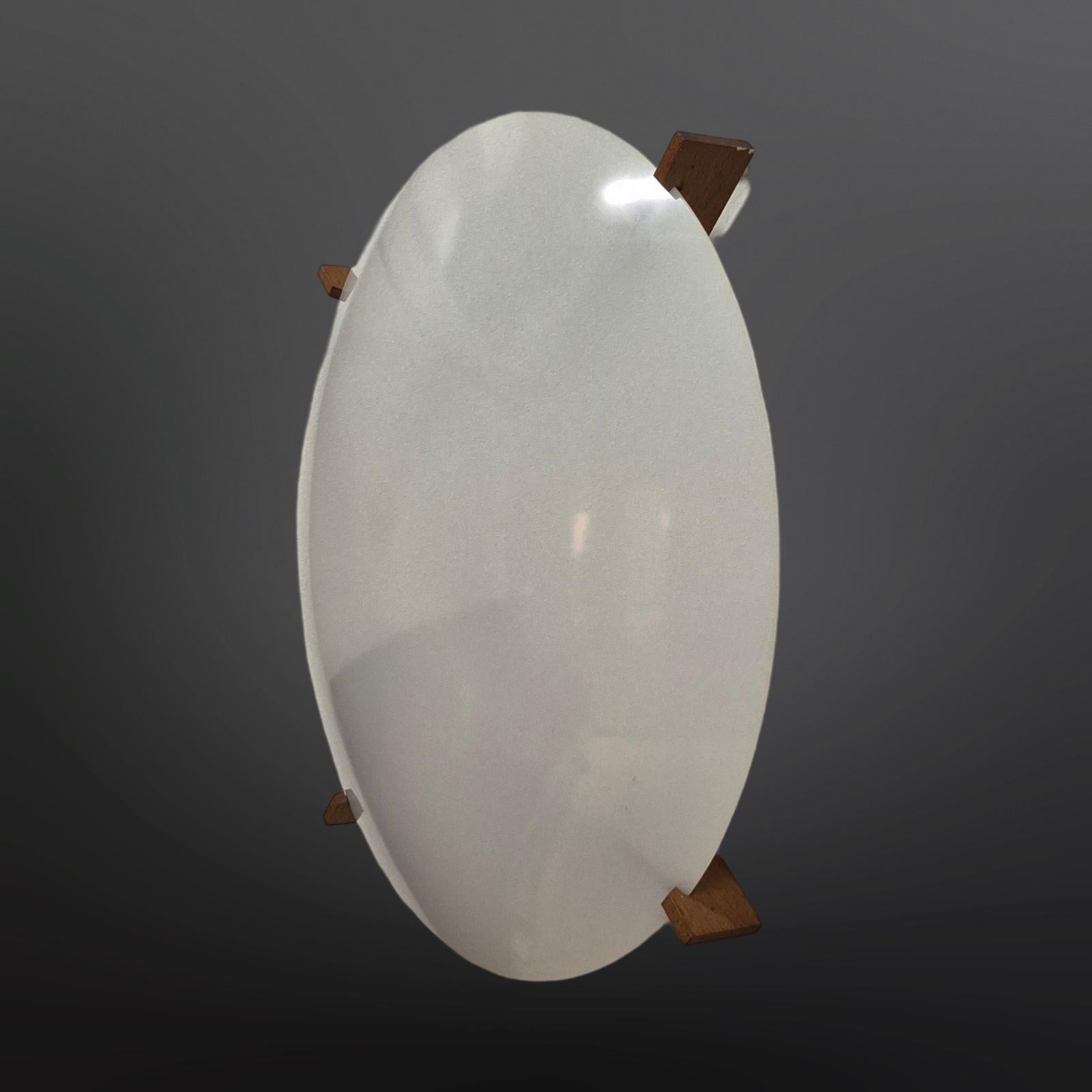 This minimalist design lamp was designd by Uno and Östen Kristiansson for the Swedish company Luxus in the 1960s. The natural teak frame holds the round white opaline plastic shade. This minimal wall or ceiling lamp is in very good vintage