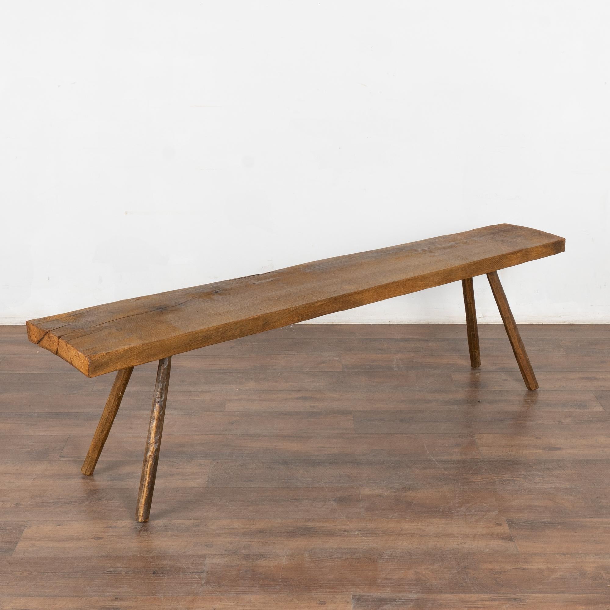 Rustic plank top bench with splay peg legs just over 6' long.
The top reveals generations of use with cracks, nicks, stains ends which add character to the narrow bench. The slightly uneven ends reveal it was hand-hewn and likely used as a work