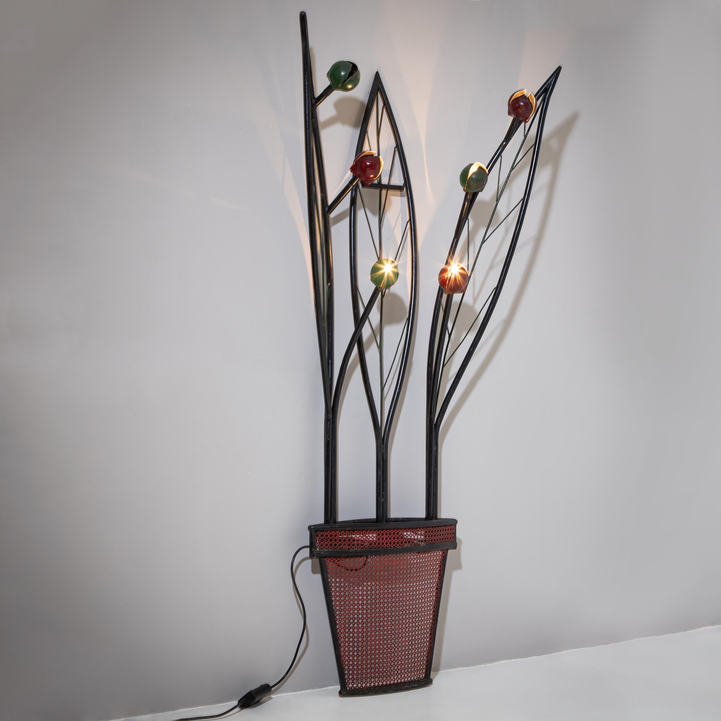 Grande lampada da terra in metallo con un disegno di piante stilizzate. Tre grandi foglie incorniciano sei prese di luce a forma di fiore, dipinte in verde e rosso. La base è disegnata come un vaso da fiori rosso con motivo a reticolo. La vernice