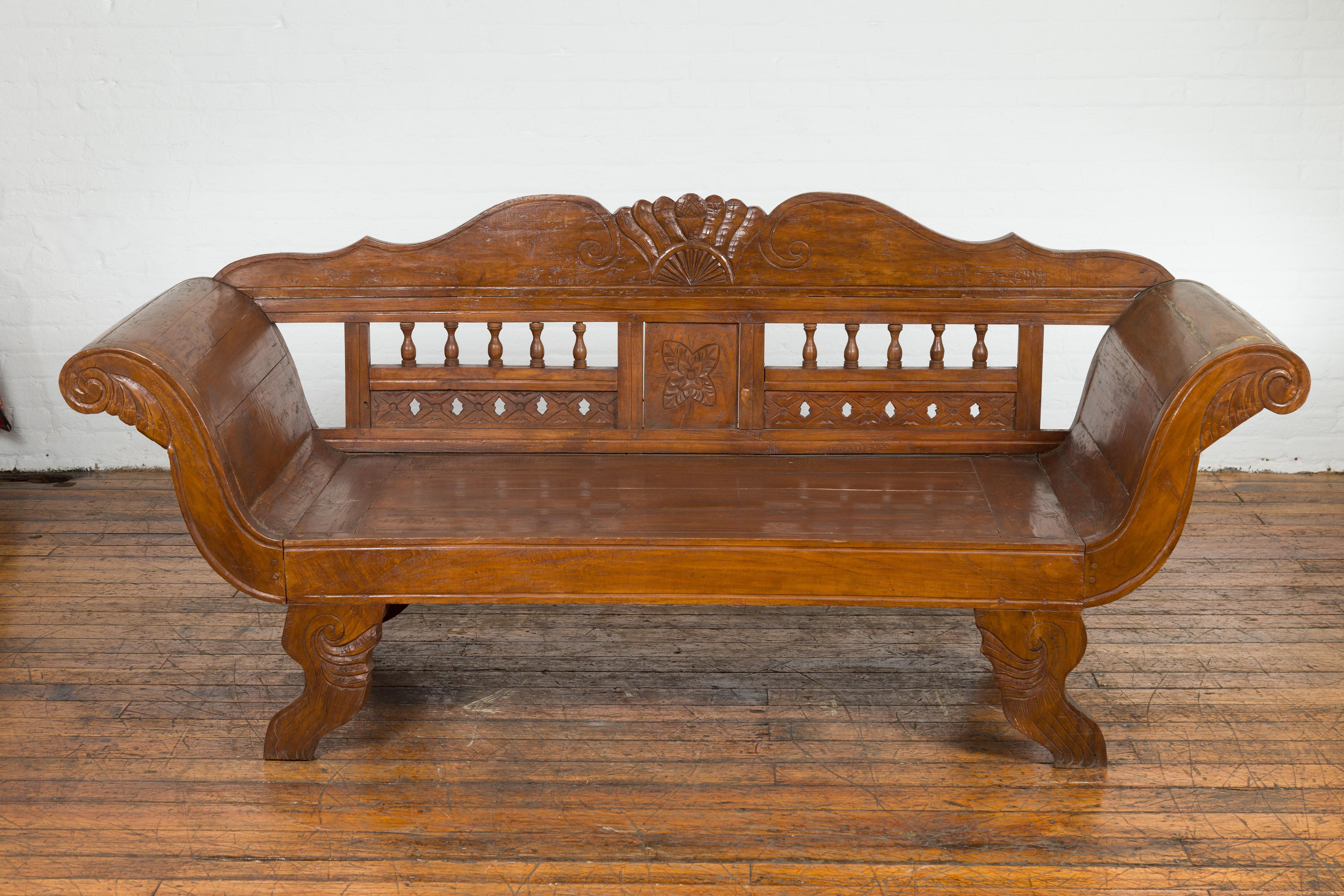An antique Javanese teak wood plantation settee from the early 20th century, with carved back, large out-scrolling arms and curving legs. Created on the Island of Java during the early years of the 20th century, this teak wood settee attracts our