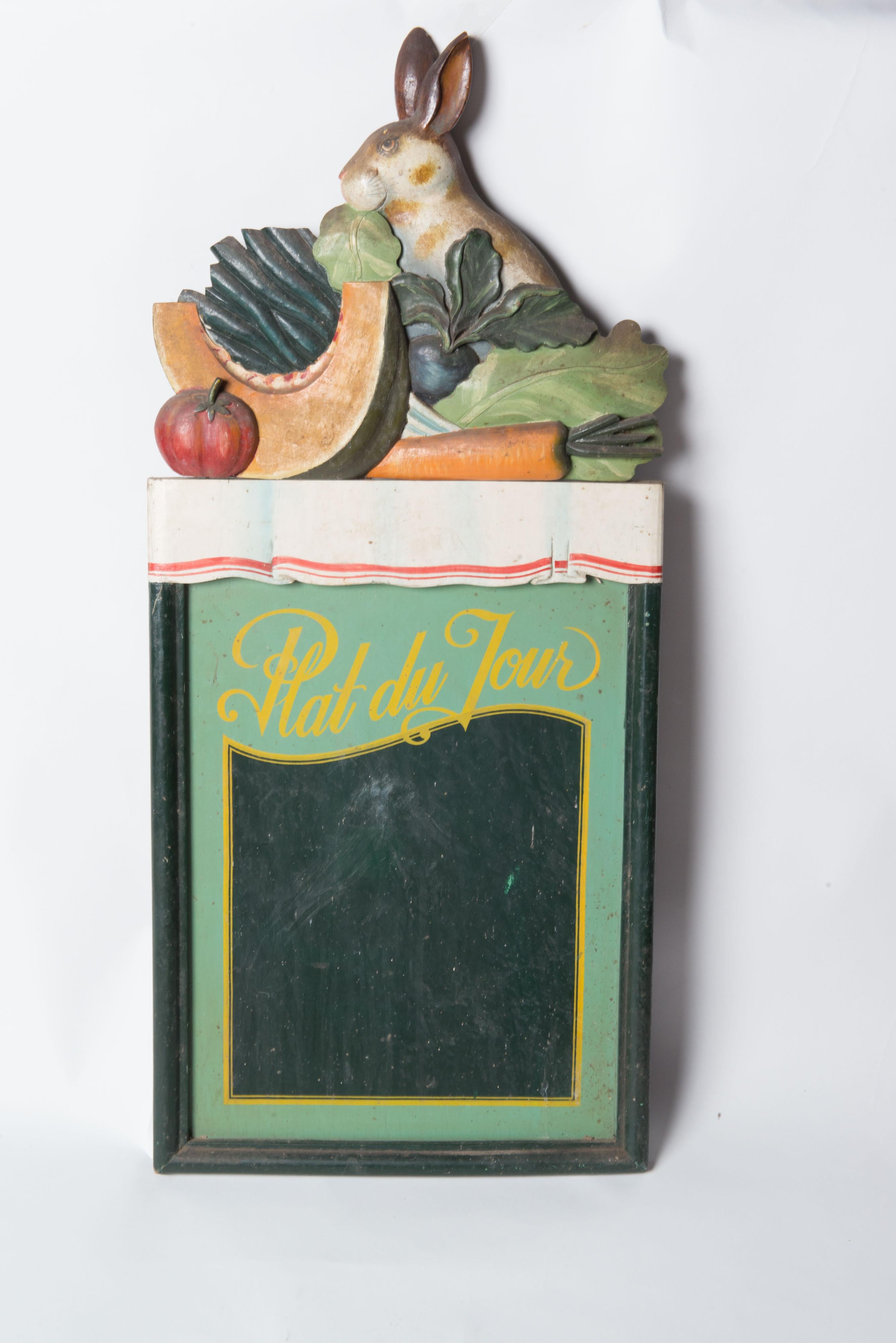 A charming Plat du Jour Rabbit chalk board. The rabbit sits with a cluster of colorful fruits and vegetable atop a tablecloth covered table all above the plat du jour chalk board. The sign is carved wood and chalk board. Use this charming sign to