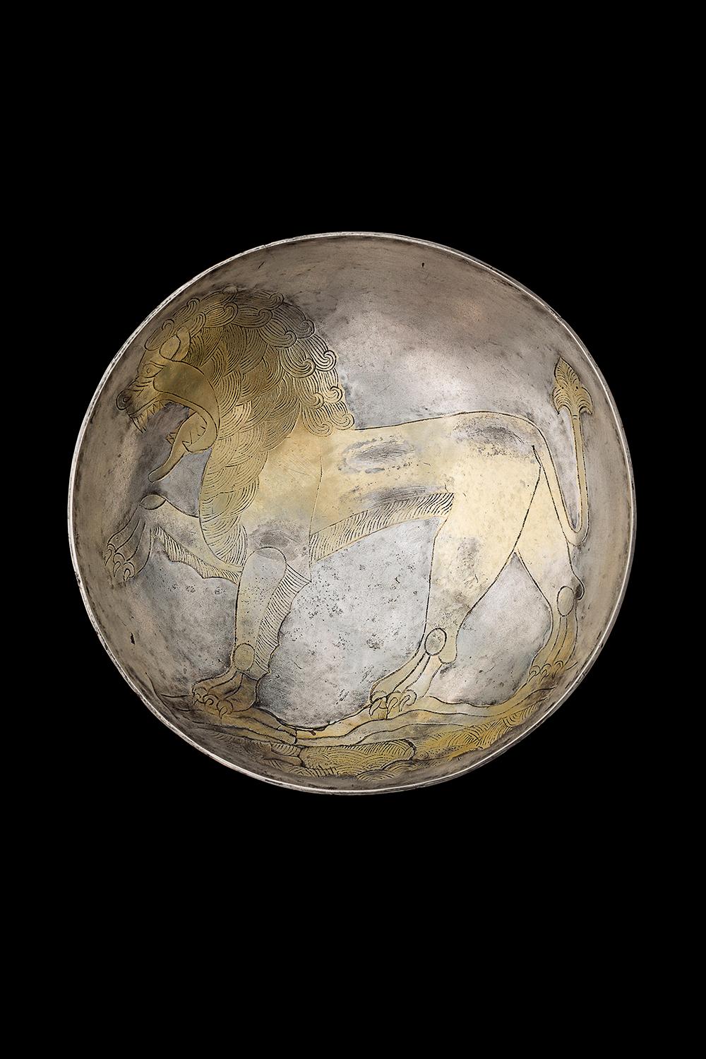 Ein Teller aus vergoldeter Silberbronze, auf dem ein großer brüllender Löwe abgebildet ist. Der Löwe ist mit erhobenem Vorderbein in der Mitte seines Weges dargestellt. Das Maul des Löwen ist weit geöffnet und zeigt seine langen, spitzen Zähne und