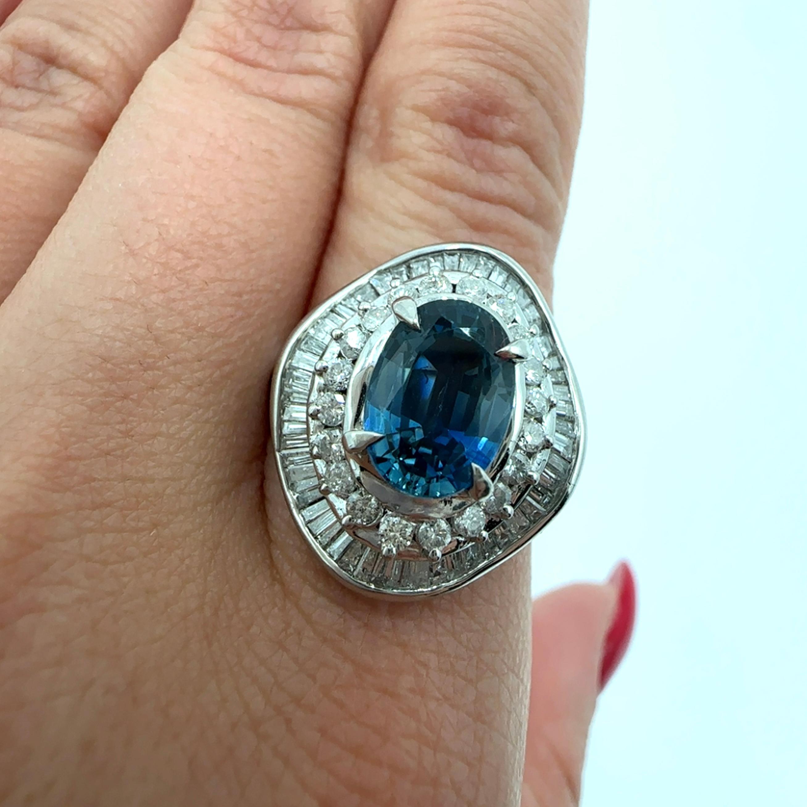 An impressive natural sapphire and diamond cocktail ring crafted in platinum 900. In the center is featured a very lively 2.76 ct, natural blue sapphire measuring 9.5mm by 6.75mm. It’s adorned by 1.05 carat total weight of natural baguette and round