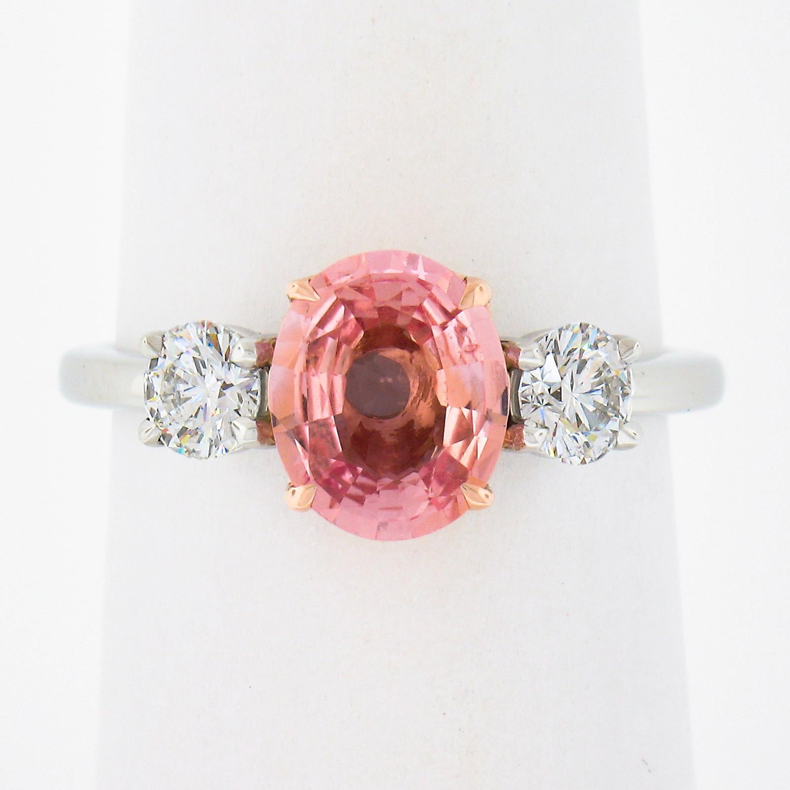 This custom and newly crafted three stone engagement ring features a gorgeous and absolutely fiery Padparadscha sapphire that is neatly prong set in the center rose gold basket which helps enhances its unusual and soothing pink-orange color. It is