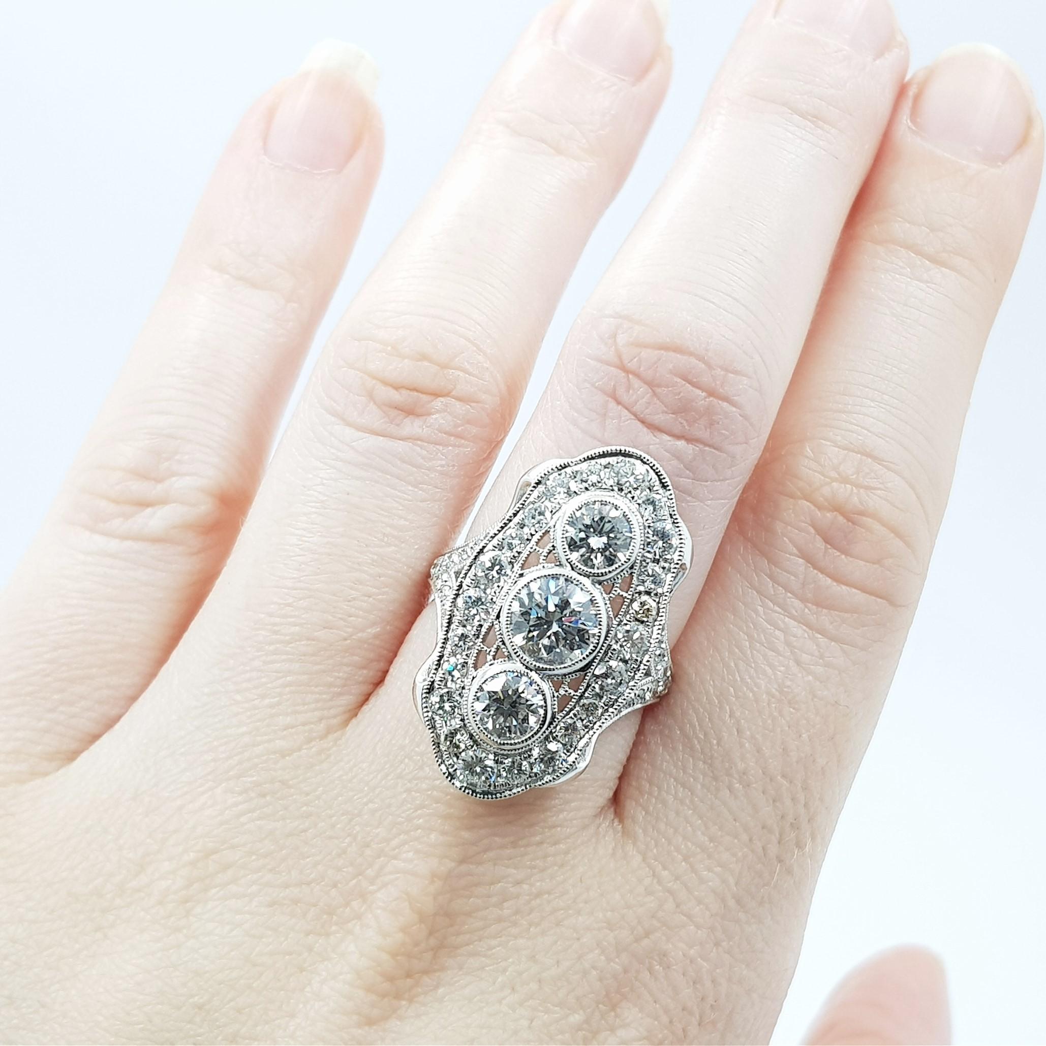 This stunning ring radiates light and calls forth the charm and elegance of vintage designs. The main trilogy of diamonds are set in individual chenier collets with milgrain edges and then are surrounded by a halo of diamonds. The total diamond