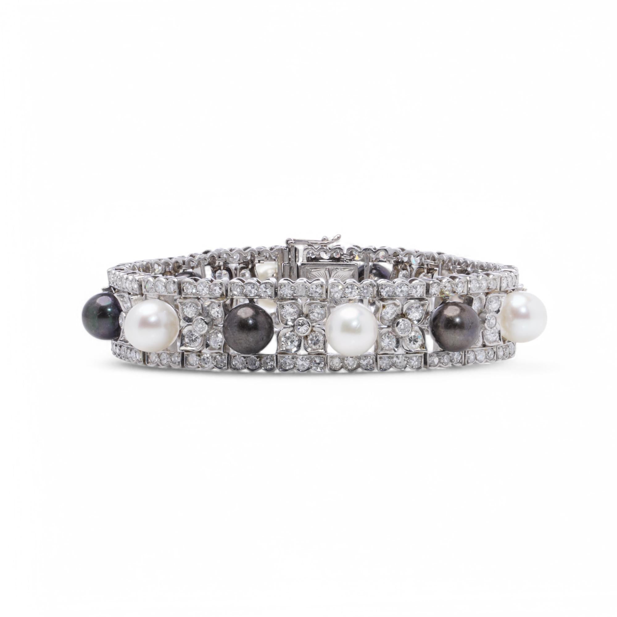 This bracelet is crafted in solid platinum, with an alternating arrangement of pearls and diamond clusters across its length. Each pearl is set within a diamond-studded floral motif, bordered by symmetrical rows of brilliant-cut diamonds. A central