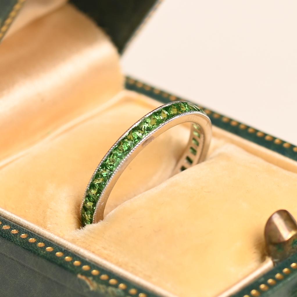 Este exquisito anillo de eternidad de platino presenta un engaste de canal sin fisuras de vibrantes granates tsavorita de talla francesa, con un total aproximado de 1,40 quilates. La banda de 2,6 mm de ancho está adornada con delicados detalles