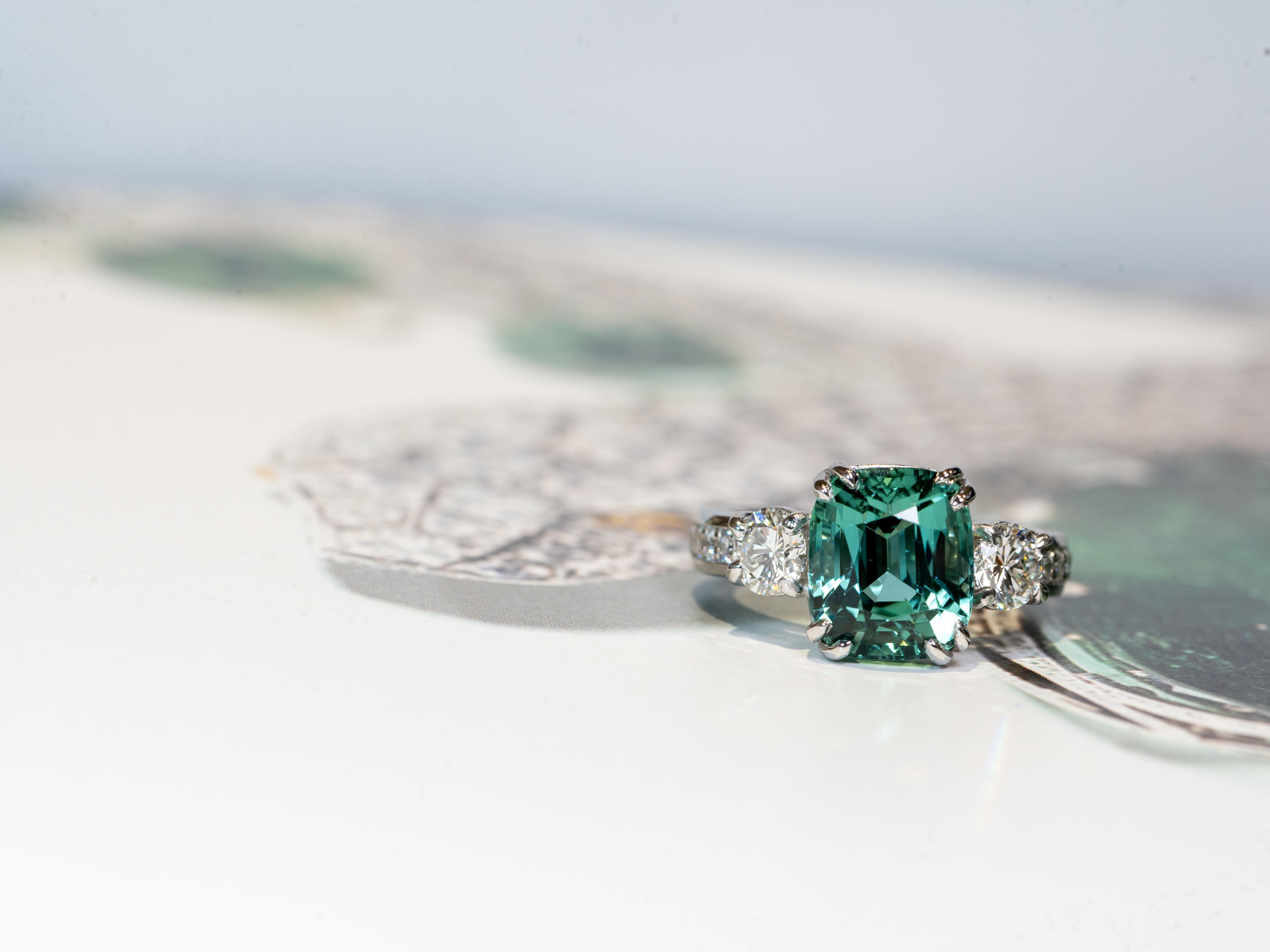 Conçue par Christopher Green, cette bague à trois pierres est fabriquée dans un peu plus de 15 grammes de platine et présente une magnifique tourmaline de couleur lagon estimée à 6-7 carats avec des dimensions de 11,8 mm x 9,4 mm. La pierre centrale