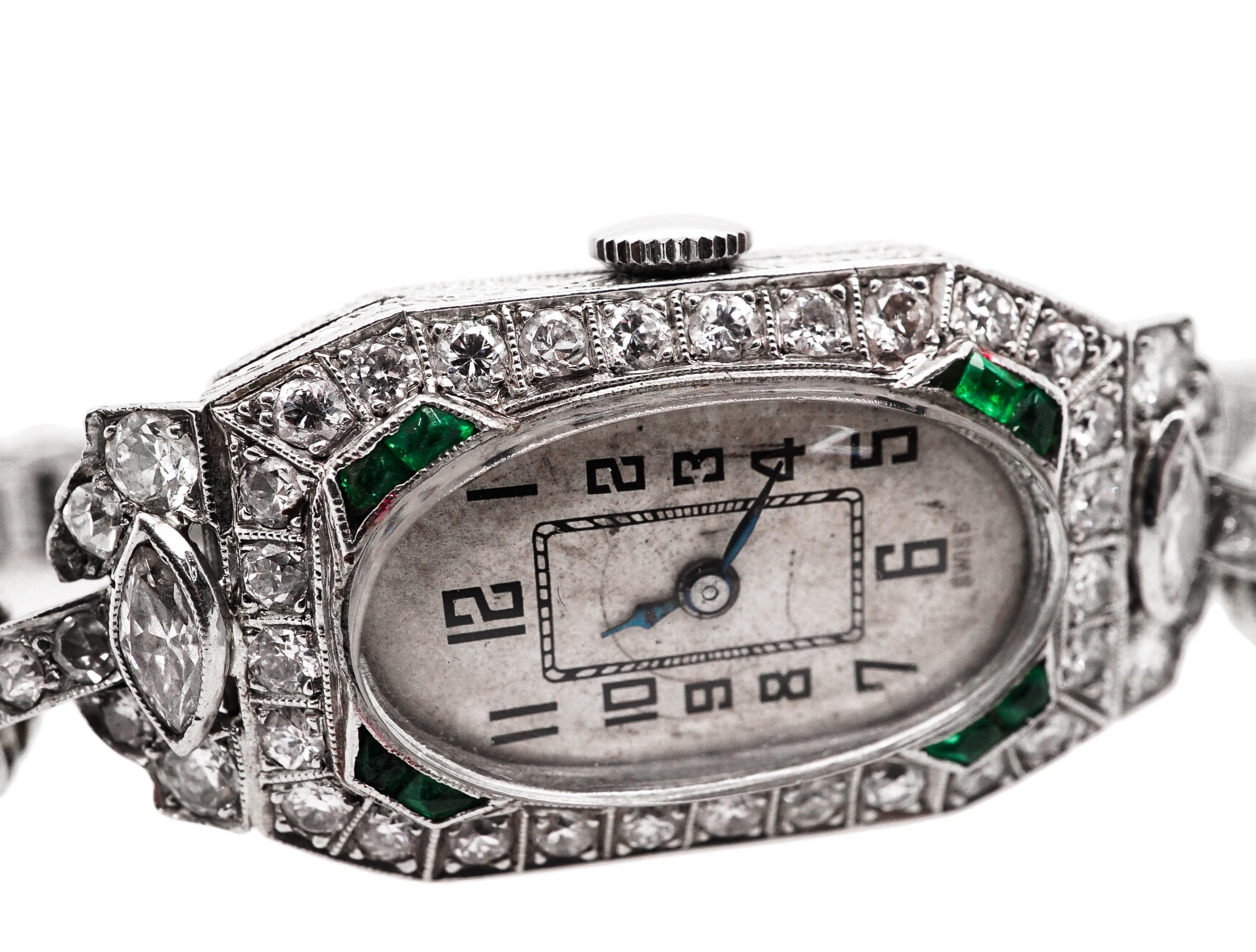 This mechanical Platinum, Diamond and Emerald Wristwatch centering an oval silver-tone dial with black Arabic numerals, within a modified rectangular diamond-set case flanked by 2 marquise-shaped diamonds, quartered by pairs of fancy-shaped
