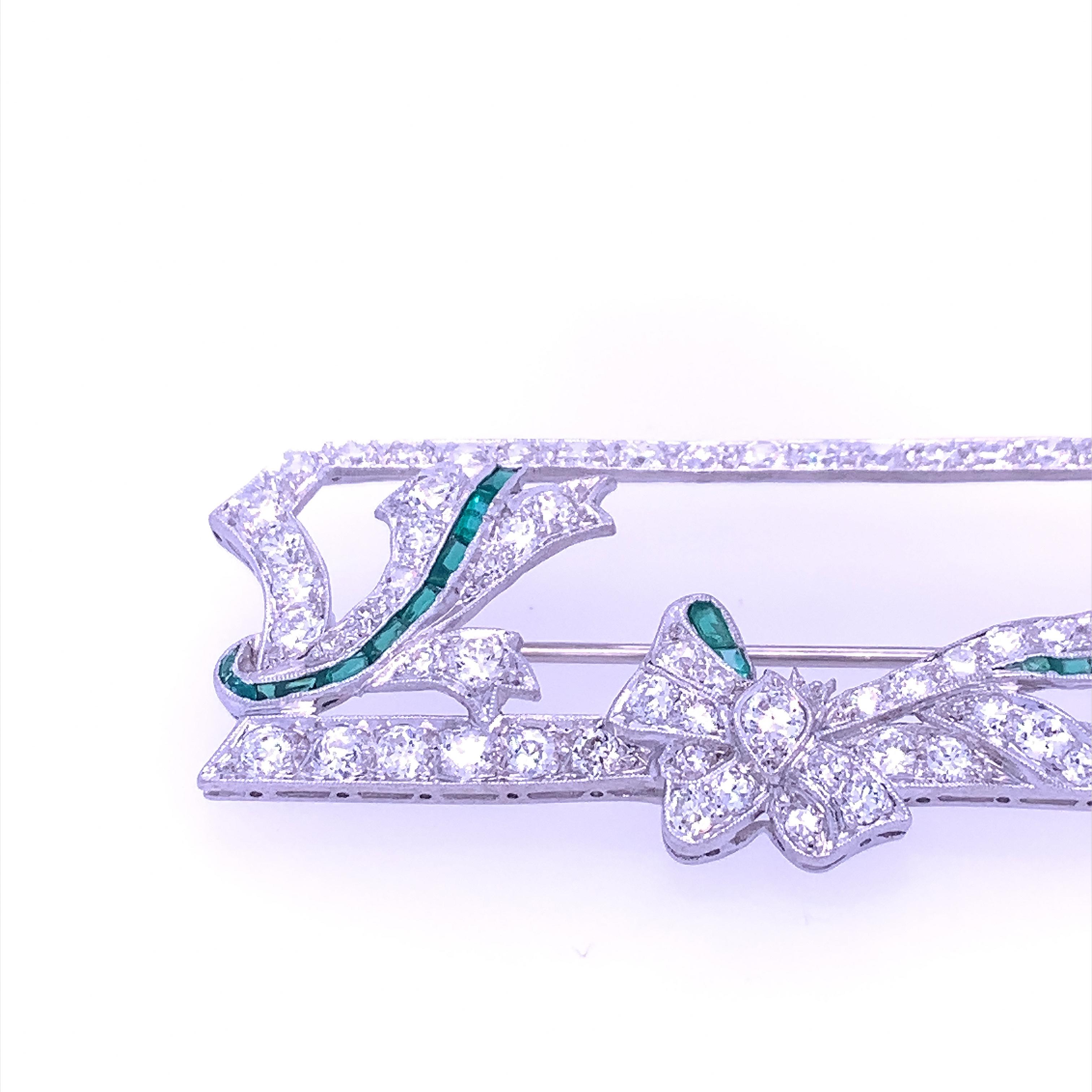 This exquisite Art Deco brooch features a diamond encrusted frame adorned with a diamond and emerald bow. A matching diamond and emerald accent anchor on the opposite side of the frame. Featuring high quality diamonds, it is enhanced by hand cut