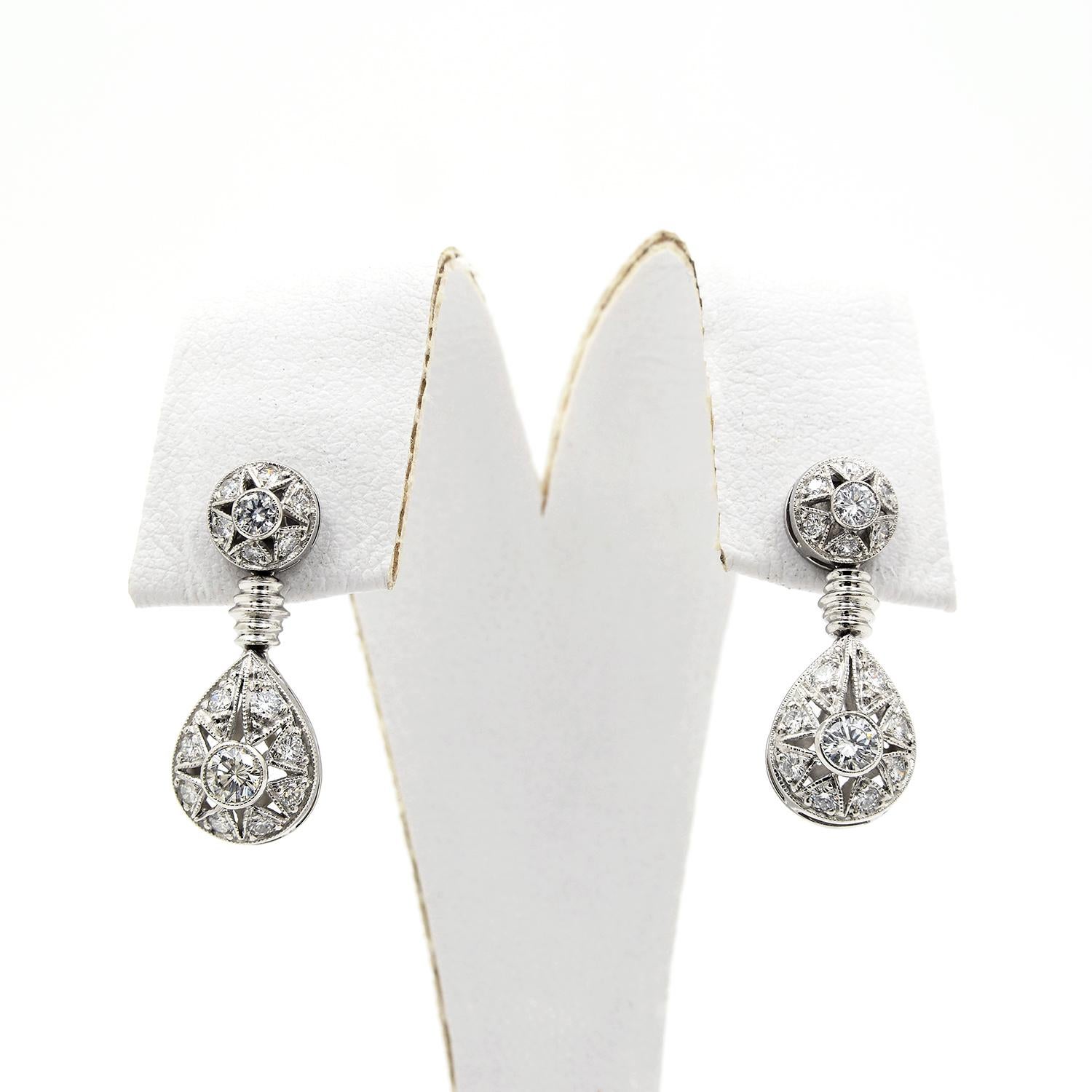 These platinum drop earrings feature a round top, bezel set with a center round brilliant cut diamond surrounded by six pave set diamonds. The drops feature a grooved bar and teardrop shape that is set with eight round brilliant cut diamonds in