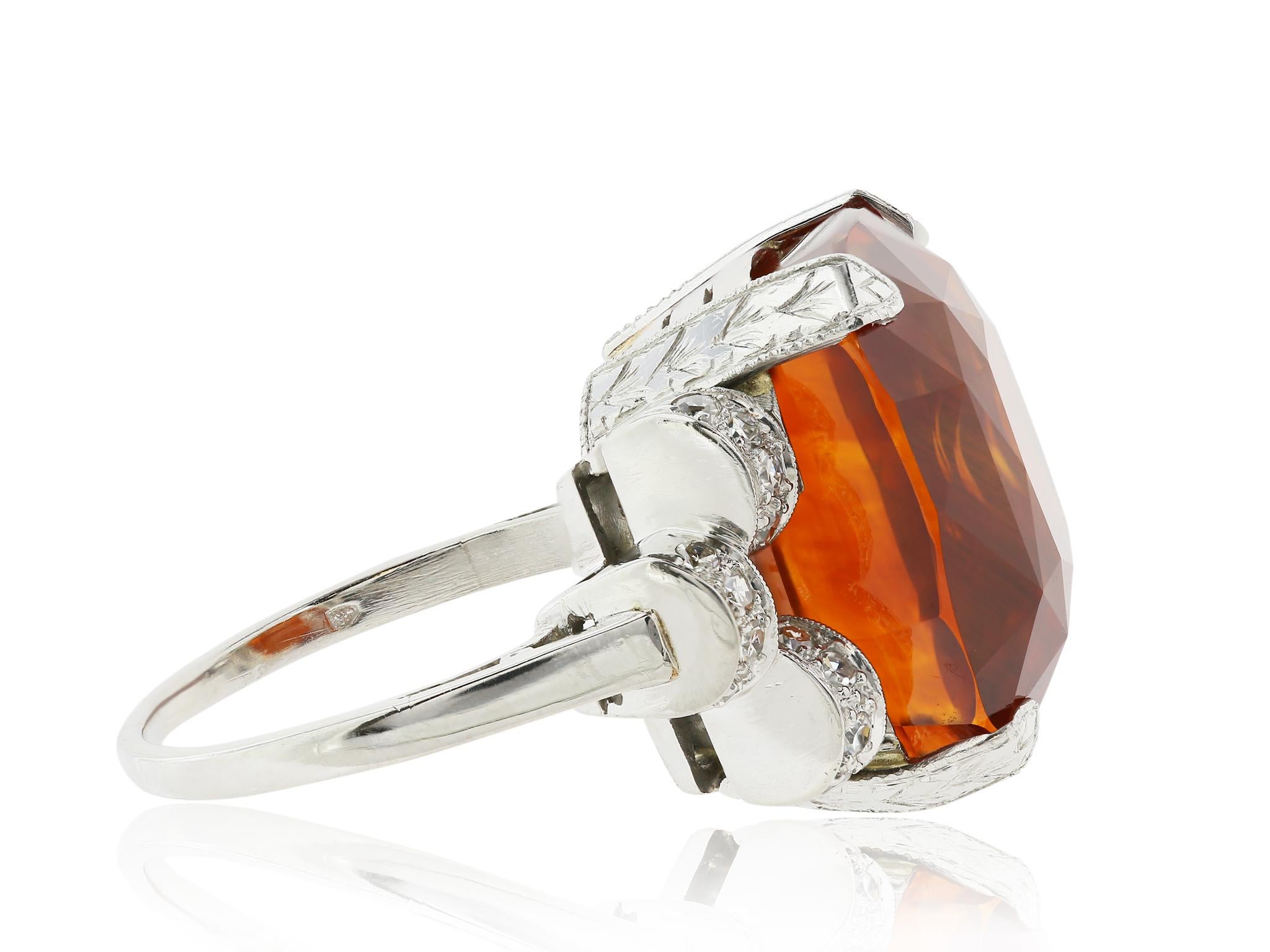 Platinum ladies ring with milgrain detail featuring one four prong-set 25.56 carat cushion modified brilliant-cut orange sapphire, flanked on either side by twelve bead-set single-cut diamond melee having a total approximate weight of 0.50 carat.
