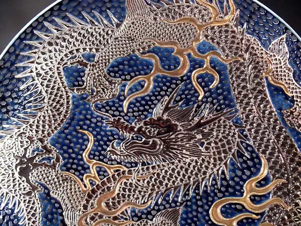 Exceptional Japanese contemporary large porcelain charger, hand painted showcasing a dramatic scene of two dragons in platinum and gold set against a stunning dimpled deep blue background, a signed masterpiece from the signature dragon collection by