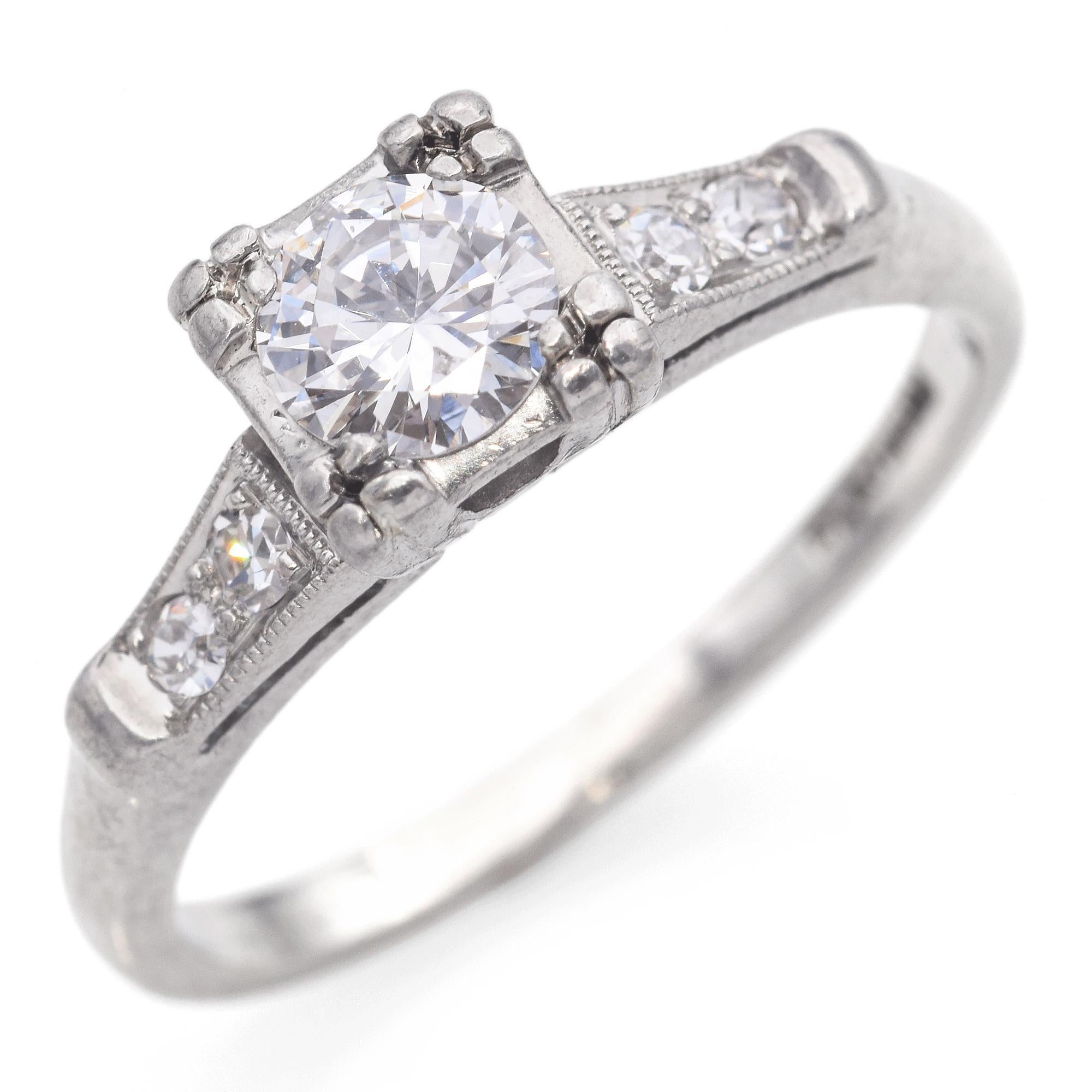 Elegamment réalisée en platine, cette bague exquise est ornée d'un diamant rond central de taille brillant, solidement maintenu par une monture carrée distinctive aux détails complexes. La pierre centrale est flanquée de deux diamants ronds plus