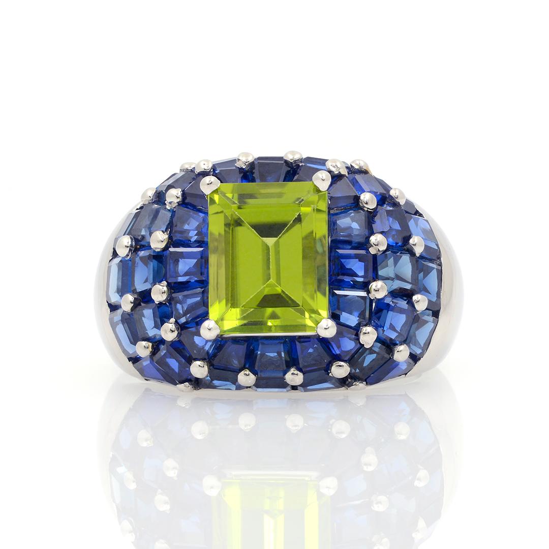 This platinum Oscar Heyman domed ring features an approximately 3.46 carat emerald cut peridot at its center surrounded by 42 calibre set blue sapphires weighing an approximate combined 10.00 carats. The ring has a polished finish and is a size