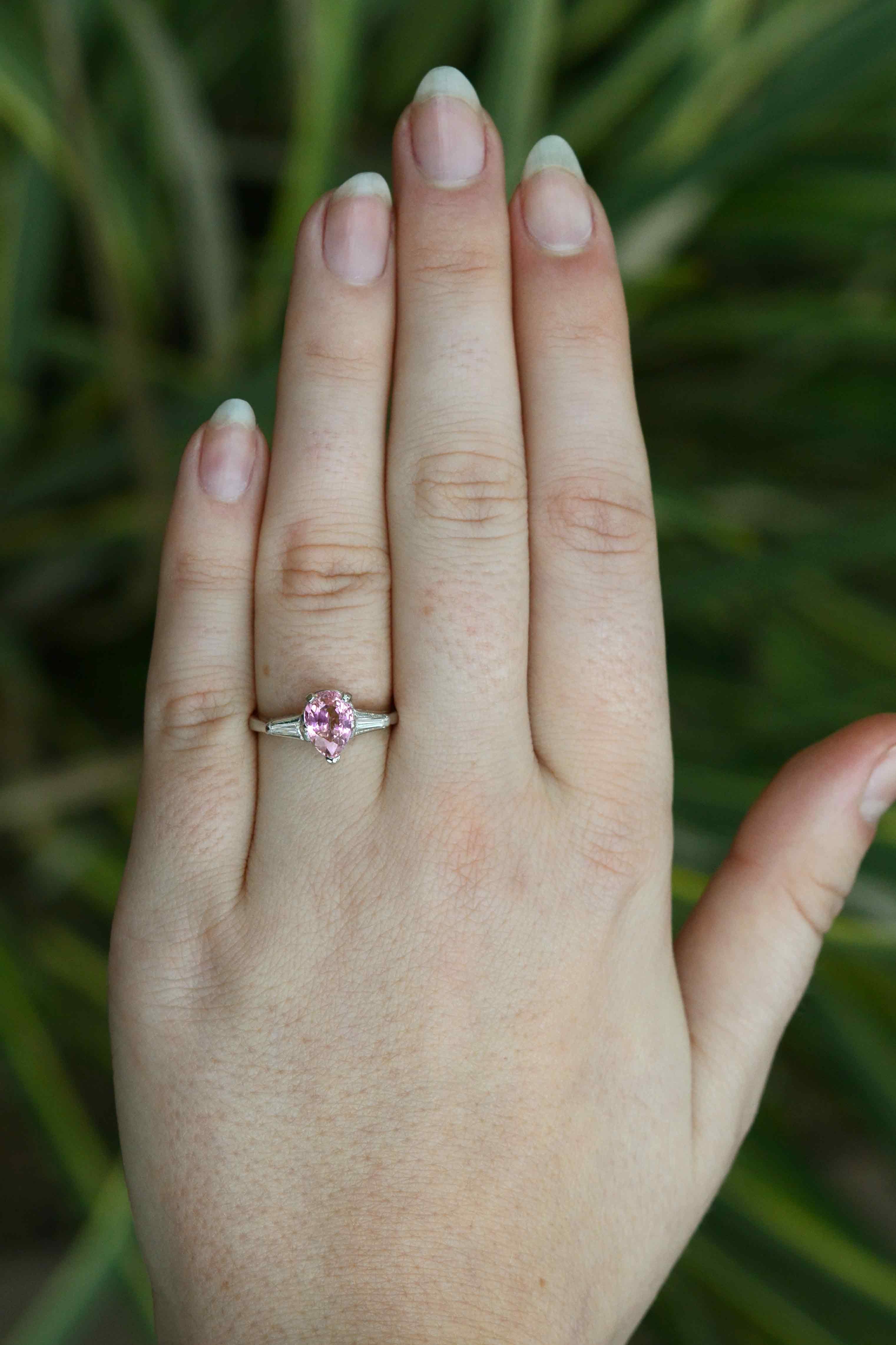 This divine pink sapphire and diamond ring makes for a perfect engagement ring. A 3 stone design representing love and compassion, the pink sapphire is lively and bright with a distinctive pear cut. The gemstone is nestled in platinum and paired