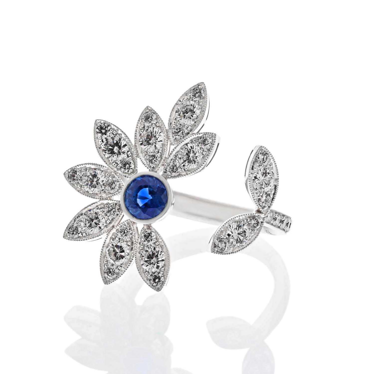 This platinum between-the-finger ring is a stunning floral design featuring a round sapphire at the center, securely set within a bezel. The petals of the flower are adorned with pave-set diamonds, adding up to an approximate total carat weight of