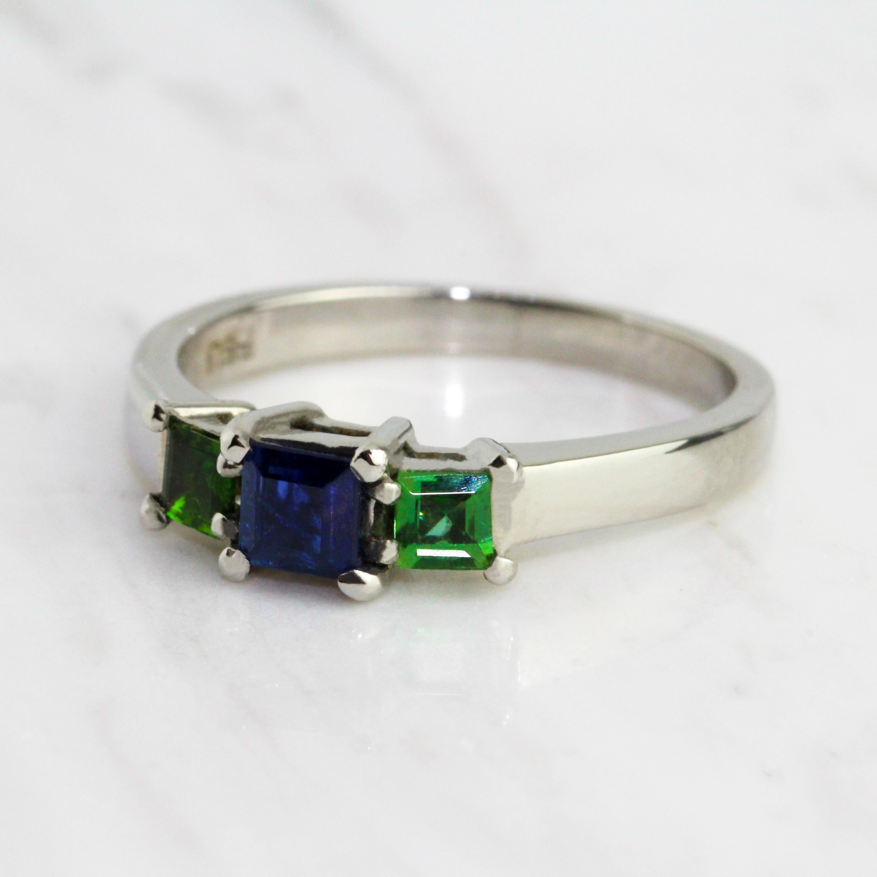 Elegant with a pop of colour, the perfect alternative engagement ring. Simple and elegant with a bold combination of colours: two square cut tsavorites frame a gorgeous square cut blue sapphire. The combination of colours and cool platinum work