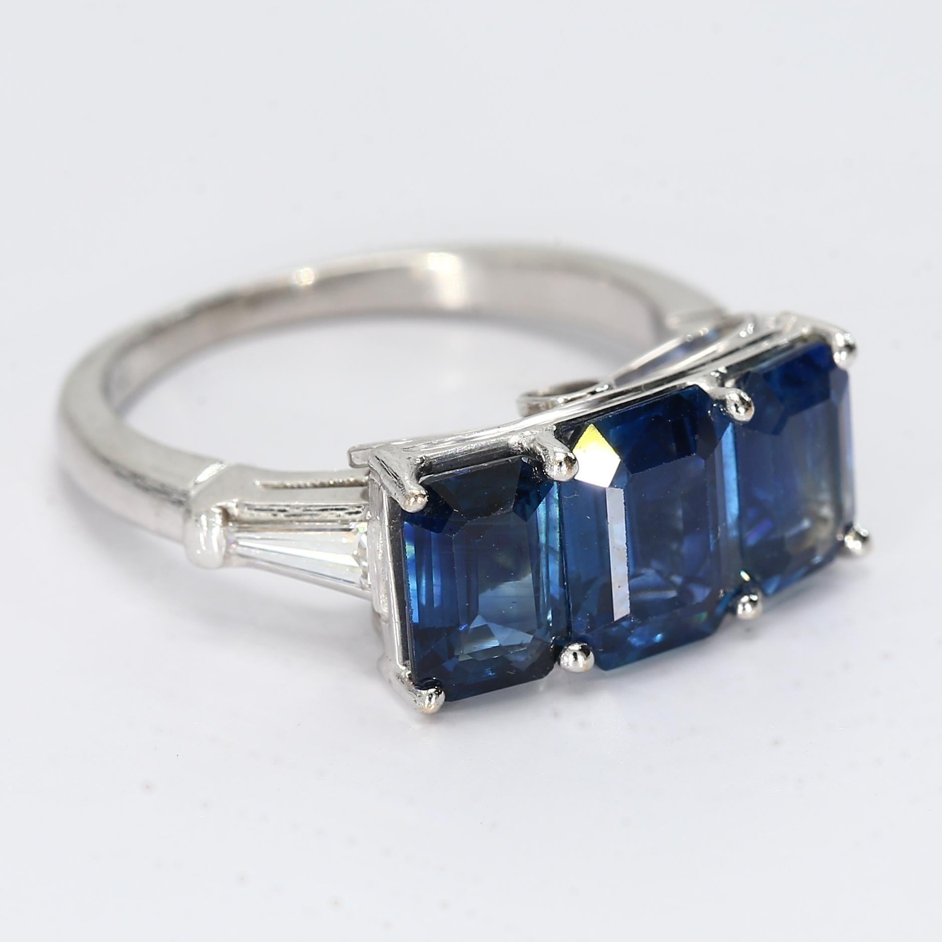This platinum three stone ring with three emerald-cut blue sapphires, and two tapered diamond baguette accent stones adorning the ring on either side.

- Stone Information -
1x - 8x6mm Emerald-Cut Blue Sapphire
2x - 7x5mm Emerald-Cut Blue