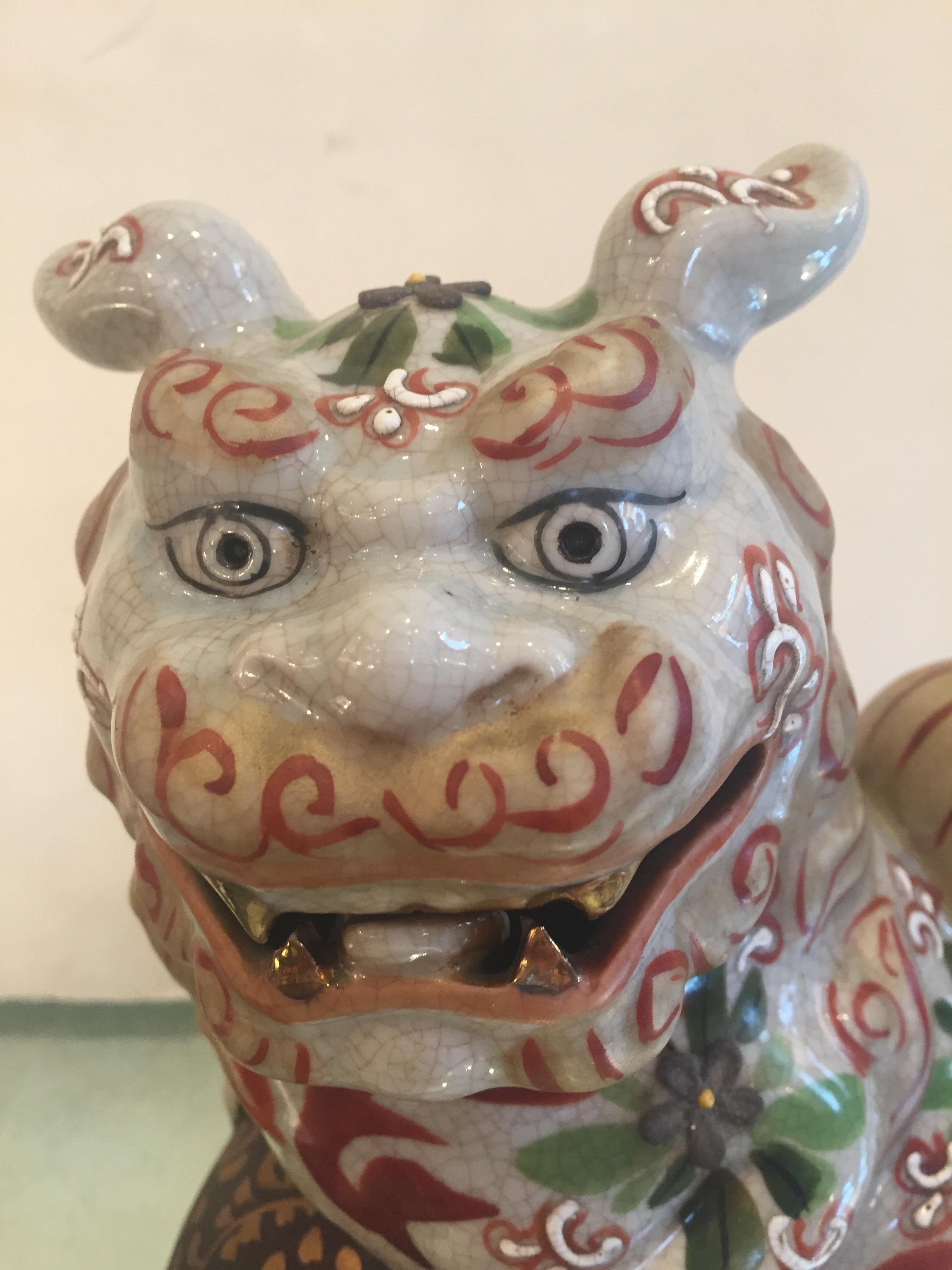 A smiling pair of decorative Chinese porcelain foo dogs having earth colors against a white background and lovely gestural poses with paws leaning on a brown and gold leaf embellished ball.