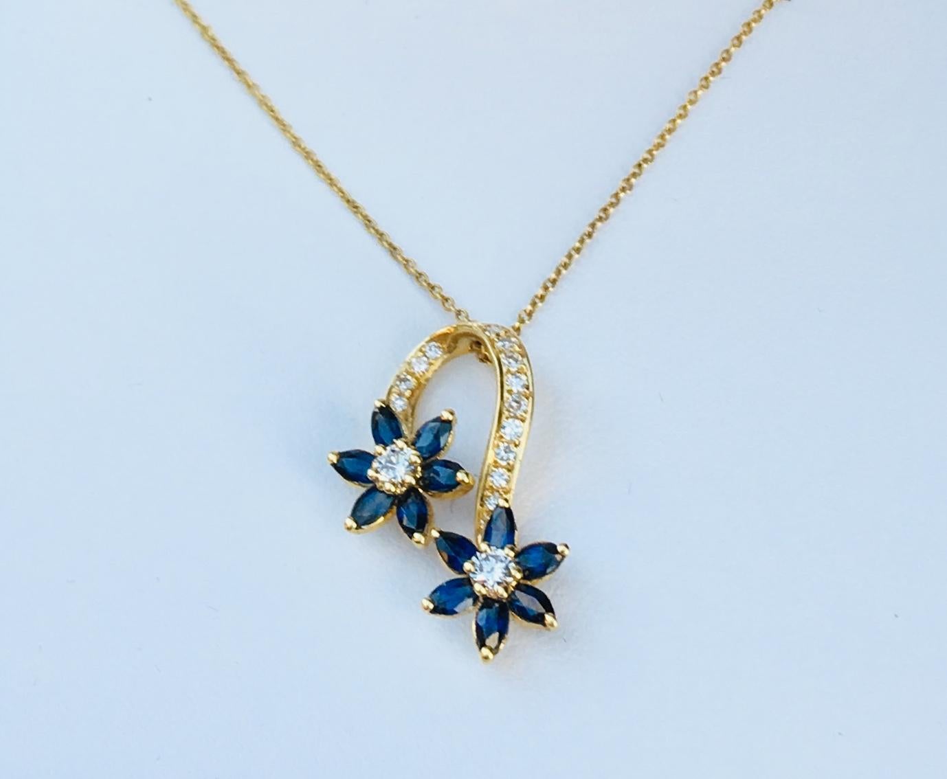 Elegant and stylized estate 18 karat yellow gold pendant on chain features two spinning flowers with prong set round brilliant diamond centers and 6 prong set, marquise cut blue sapphire petals.  Flowers are connected by a graceful 18 karat yellow