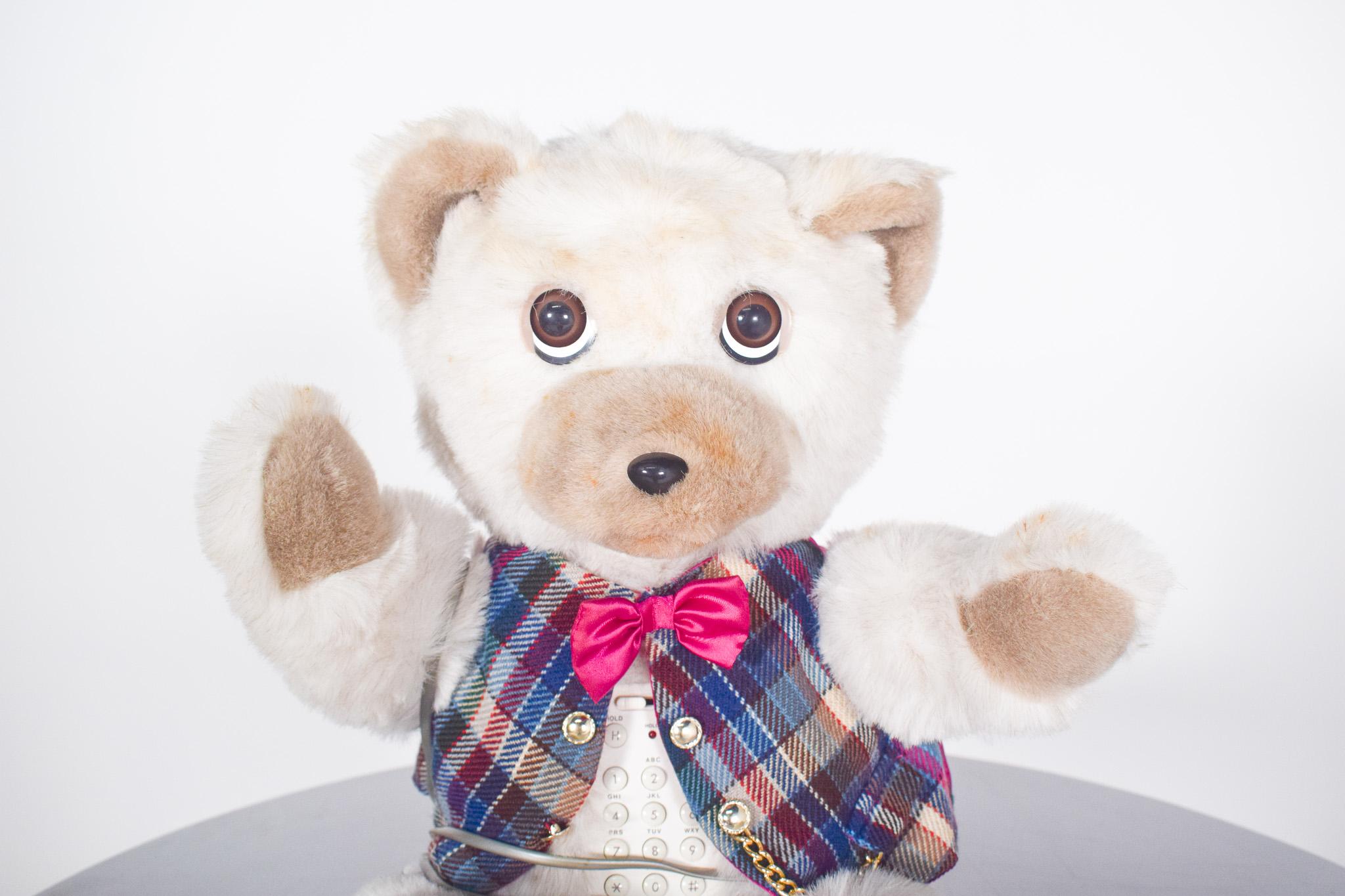 Ourson avec téléphone mains libres intégré. KC Bearifone II, produit par TeleConcepts Inc. dans les années 1990. Il s'agit d'un téléphone mains libres avec haut-parleur intégré, déguisé en poupée, avec un gilet à carreaux et un nœud papillon rose,