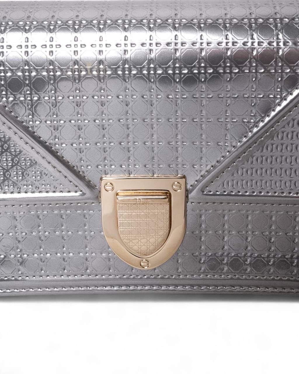 Pochette firmata Dior, modello Diorama, misura Small, realizzata in pelle verniciata nella colorazione silver con hardware dorato. Dotata di una patta frontale con chiusura a bottone, l’interno è rivestito in pelle liscia grigia e offre molteplici