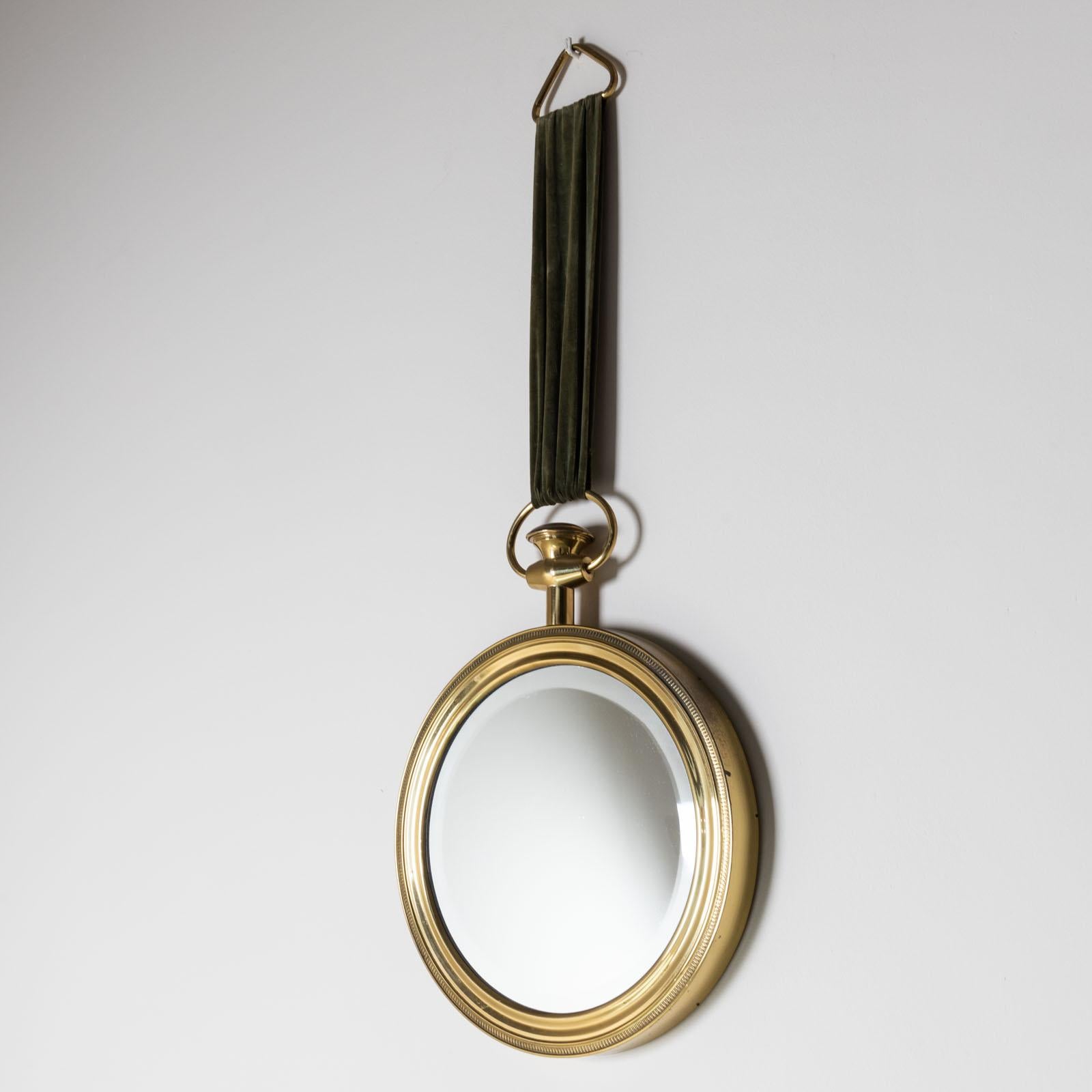 Specchio decorativo da parete a forma di orologio da tasca sovradimensionato, nello stile di Piero Fornasetti. La cornice in ottone profilato è ornata da un raffinato fregio decorativo lungo il bordo esterno. Lo specchio è sospeso da un nastro di