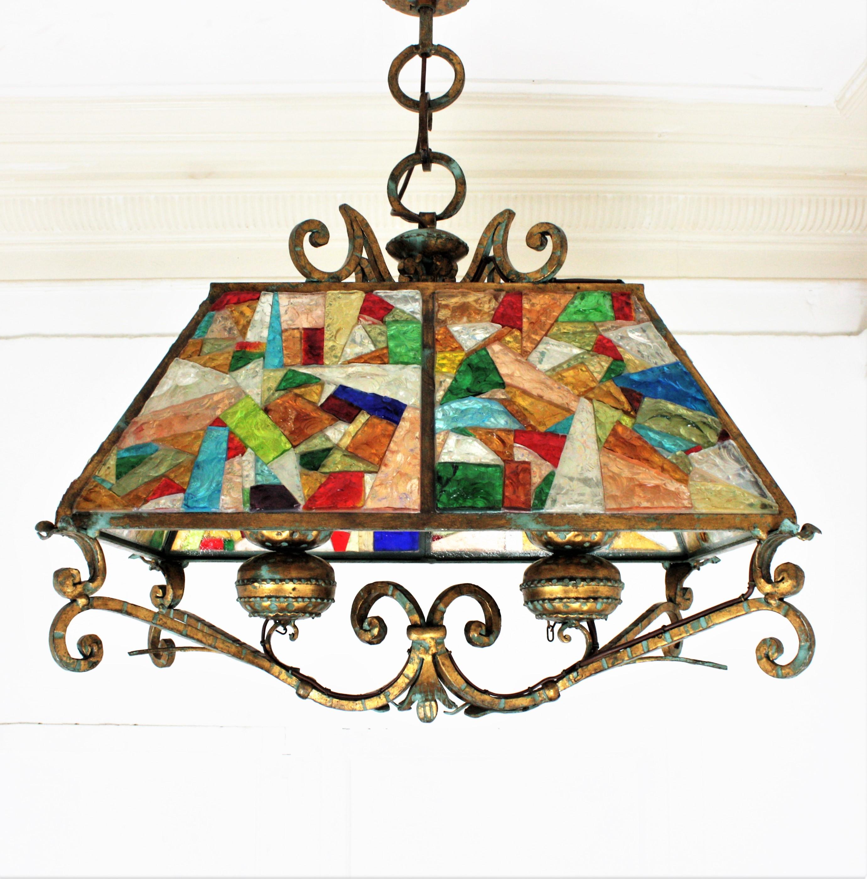 A massive multi-color hammered glass and gilt wrought iron suspension lamp. Manufactured by Longobard, the concurrent of Poliarte at the 1960s.
This ginormous lantern in Brutalist style has classical accents. The structure is made in heavy wrought