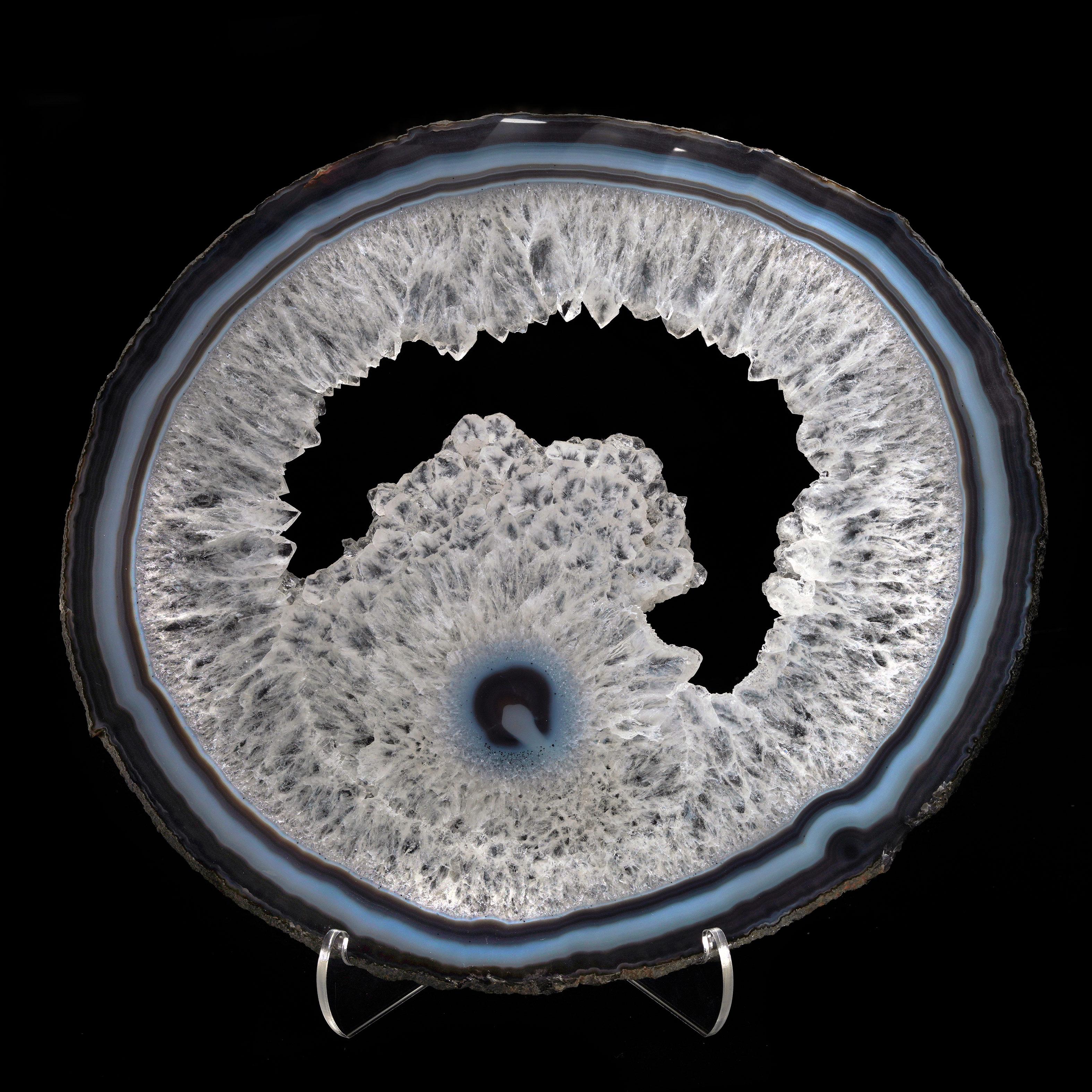 Questa splendida fetta di geode di agata lucidata proveniente dal Brasile presenta affascinanti bande concentriche di agata blu, grigia e bianca che circondano un radioso centro di quarzo cristallino. Lo scintillio naturale del quarzo aggiunge un