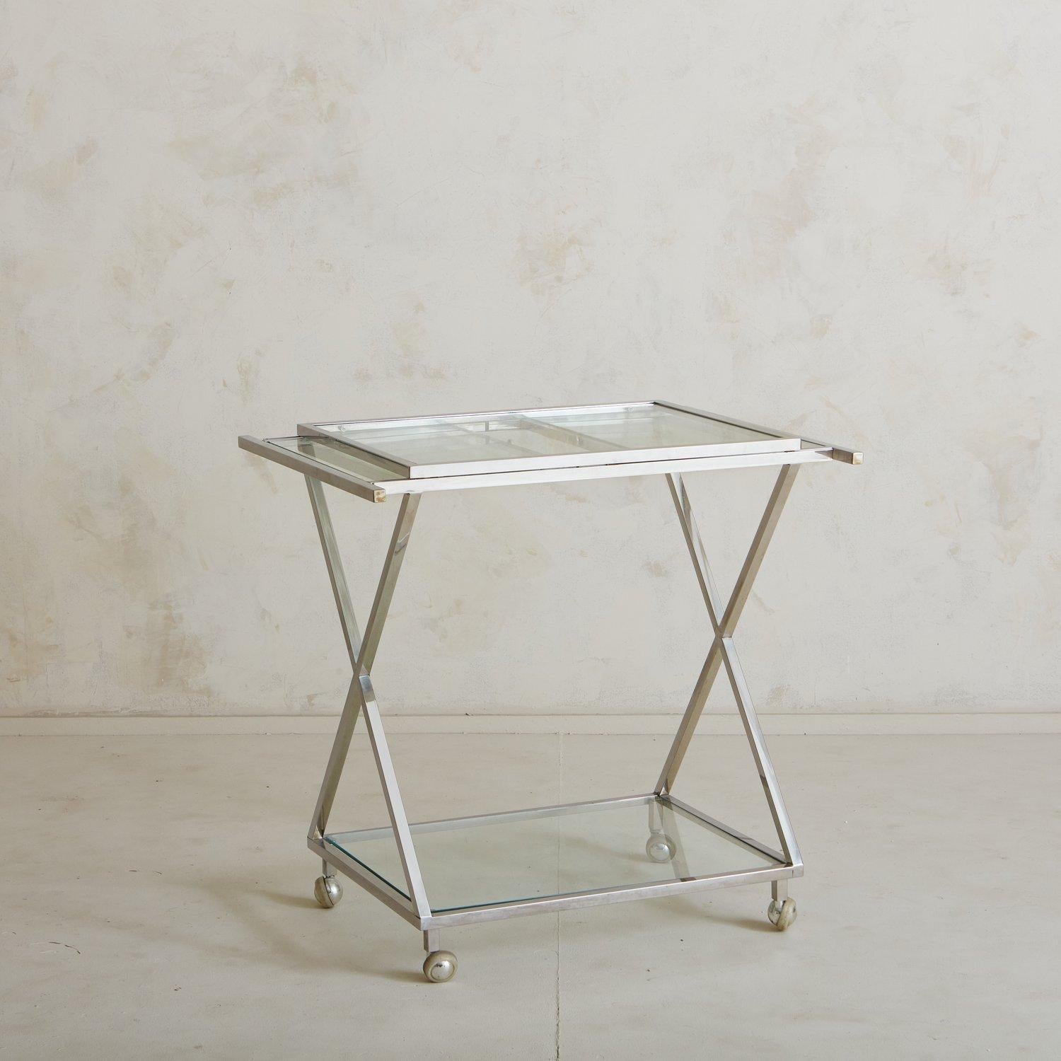 A vintage polished chrome two-tier bar cart featuring rectangular glass shelves and two extendable leaves. This piece has a sleek profile with X-detailing and stands on four casters for easy mobility. USA, 1960s.

 Measures: 31.5