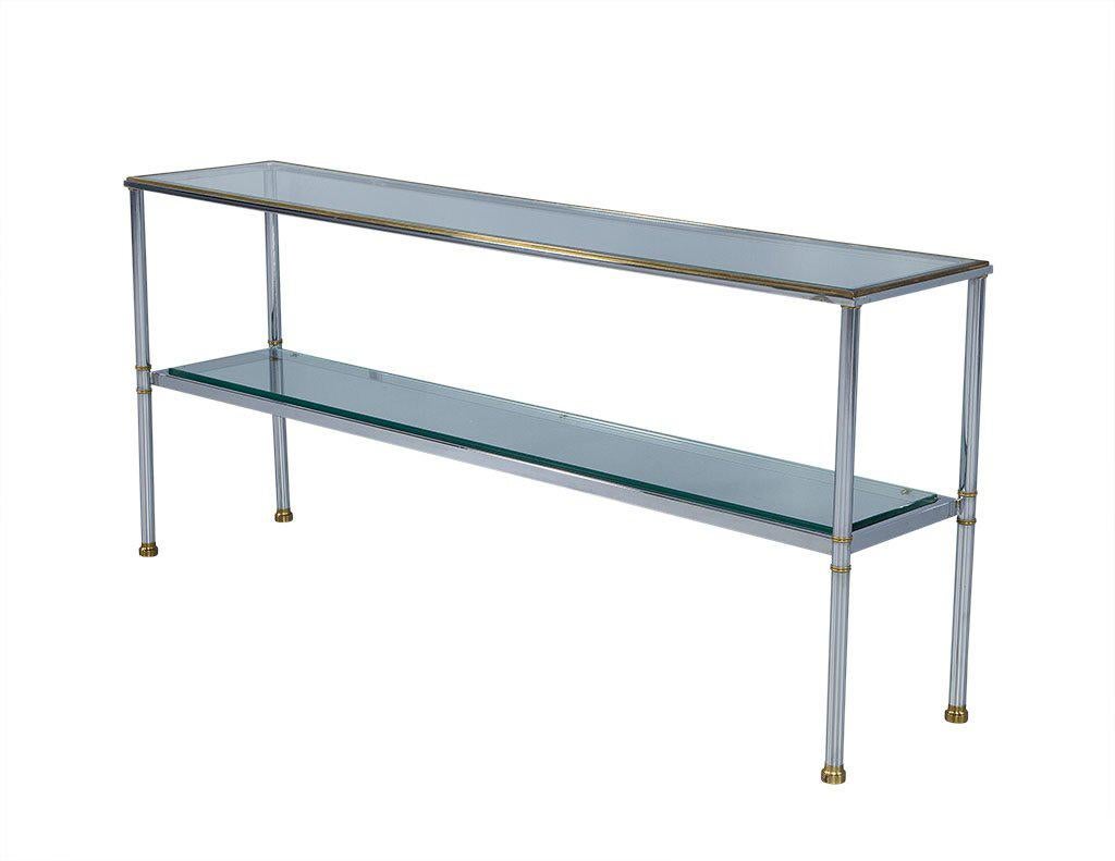 Elegant and sleek, this console table combines a clear glass plateau with a polished chrome body and brass accents. These accents are seen on the foot caps and on the trim around the top. A middle glass shelf provides extra display space while