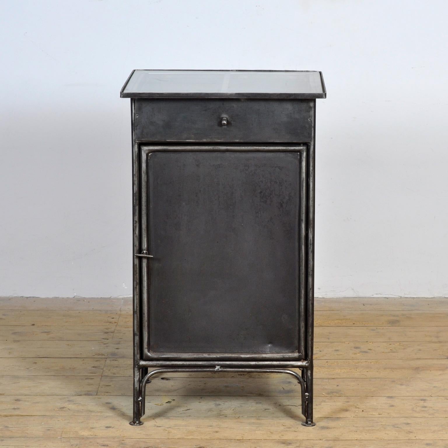 Cette table de chevet en fer provient d'un hôpital de Budapest, vers 1910. La table a été entièrement dépouillée jusqu'au métal, puis polie, ce qui met en valeur son aspect industriel. Le plateau en verre apporte une touche élégante et légère, ce