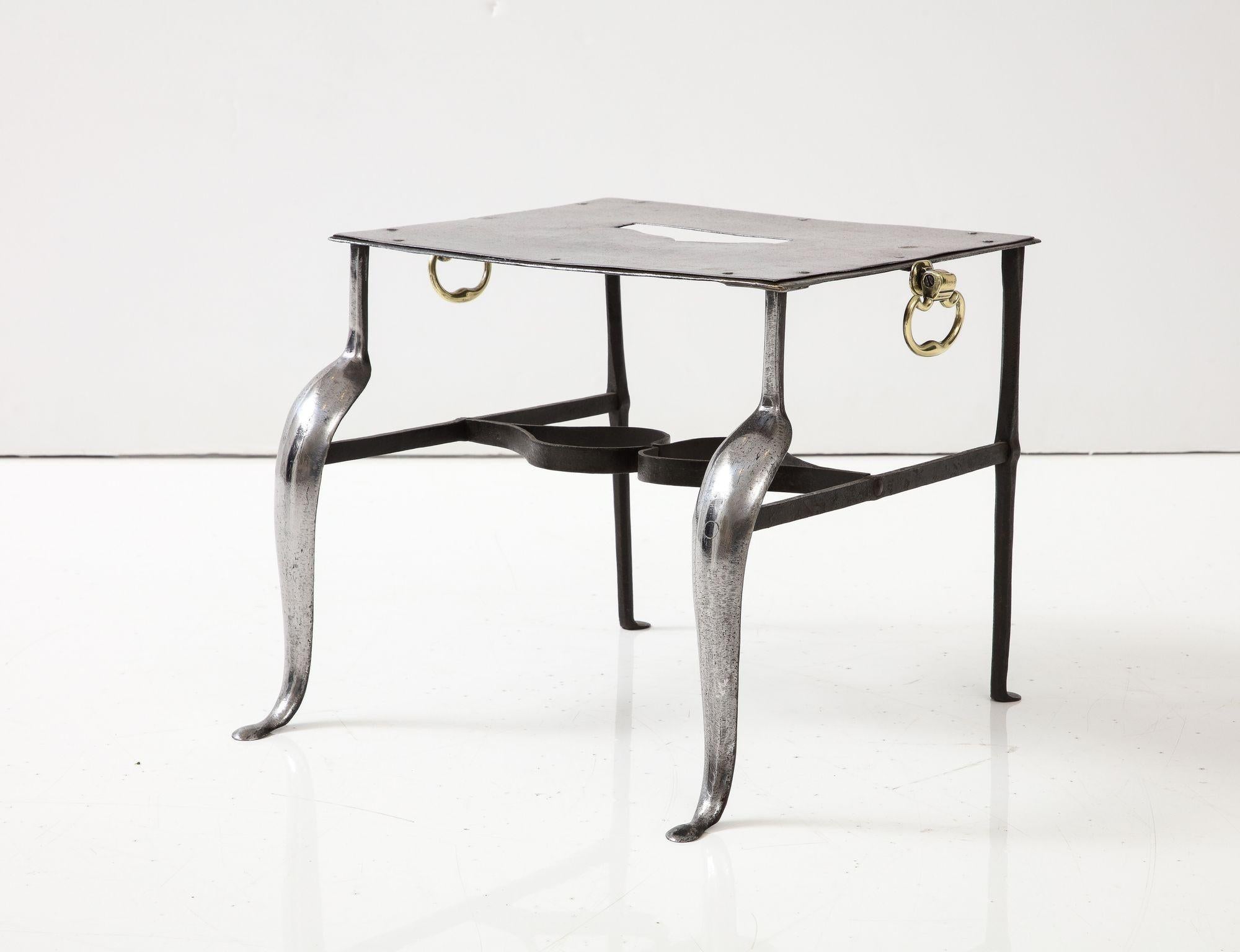 A polished steel stand originally used to hold hot kettles and pots next to a fireplace and known as a trivet.  Rectangular polished top with ogee cutout handle on polished steel cabriole front legs with wrought iron H stretcher featuring double