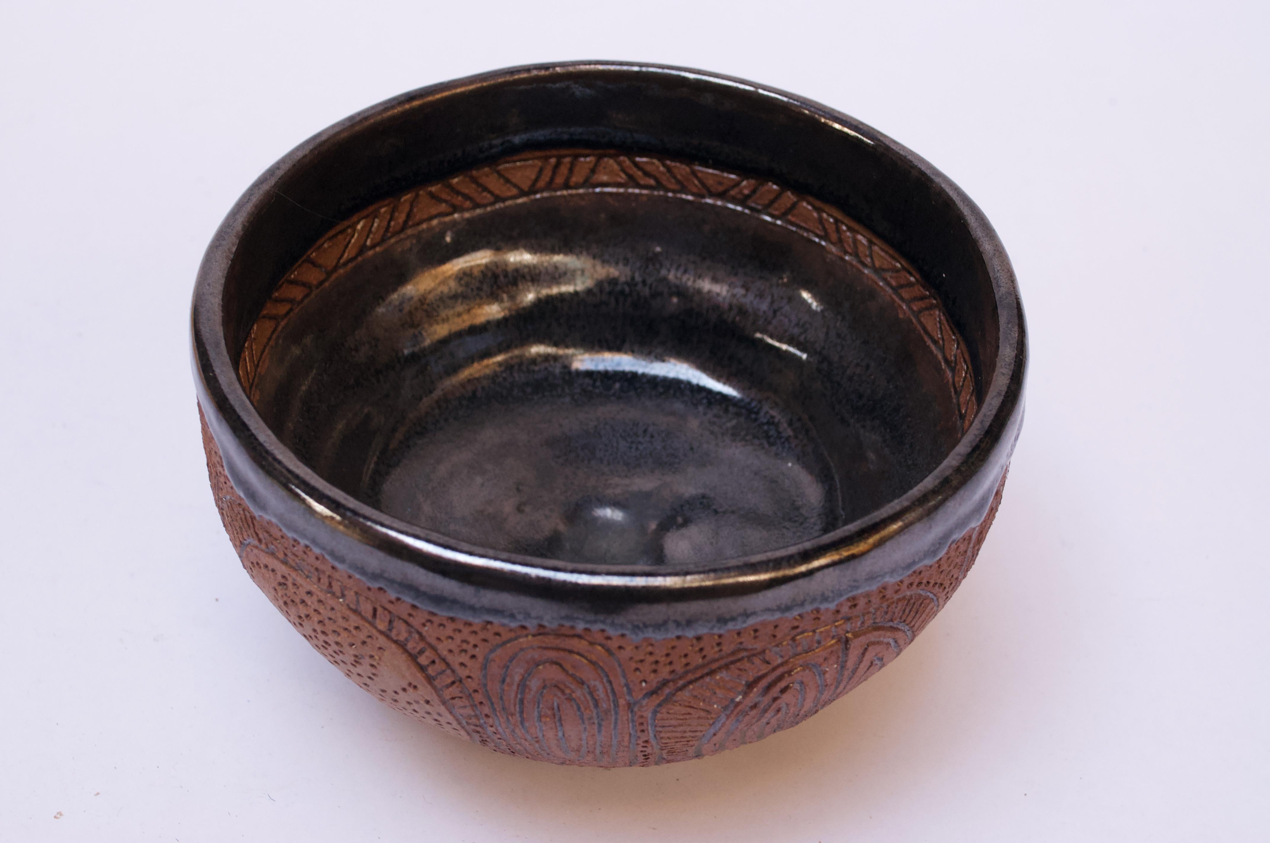 Heavily textured footed bowl with sgraffito and pinhole decoration throughout. Black interior glaze has a subtle metallic sheen and features a thin circular band with incised triangular and linear detail. 
Signed 