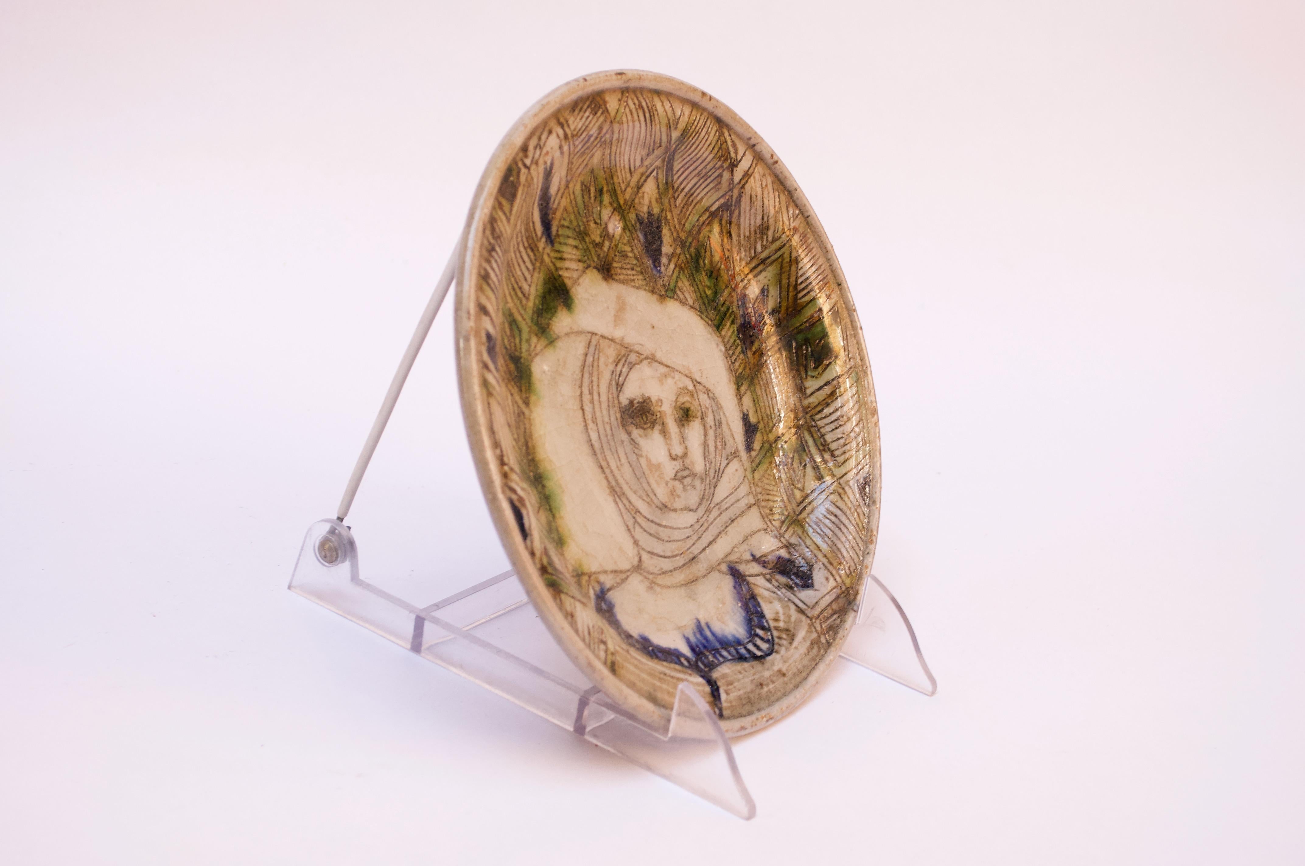 Studio ceramic decorative plate depicting a woman with a headscarf. Linear and geometric details present along with pops of blue and green to enhance the smattering of floral decorations around the central figure. 
Signed 