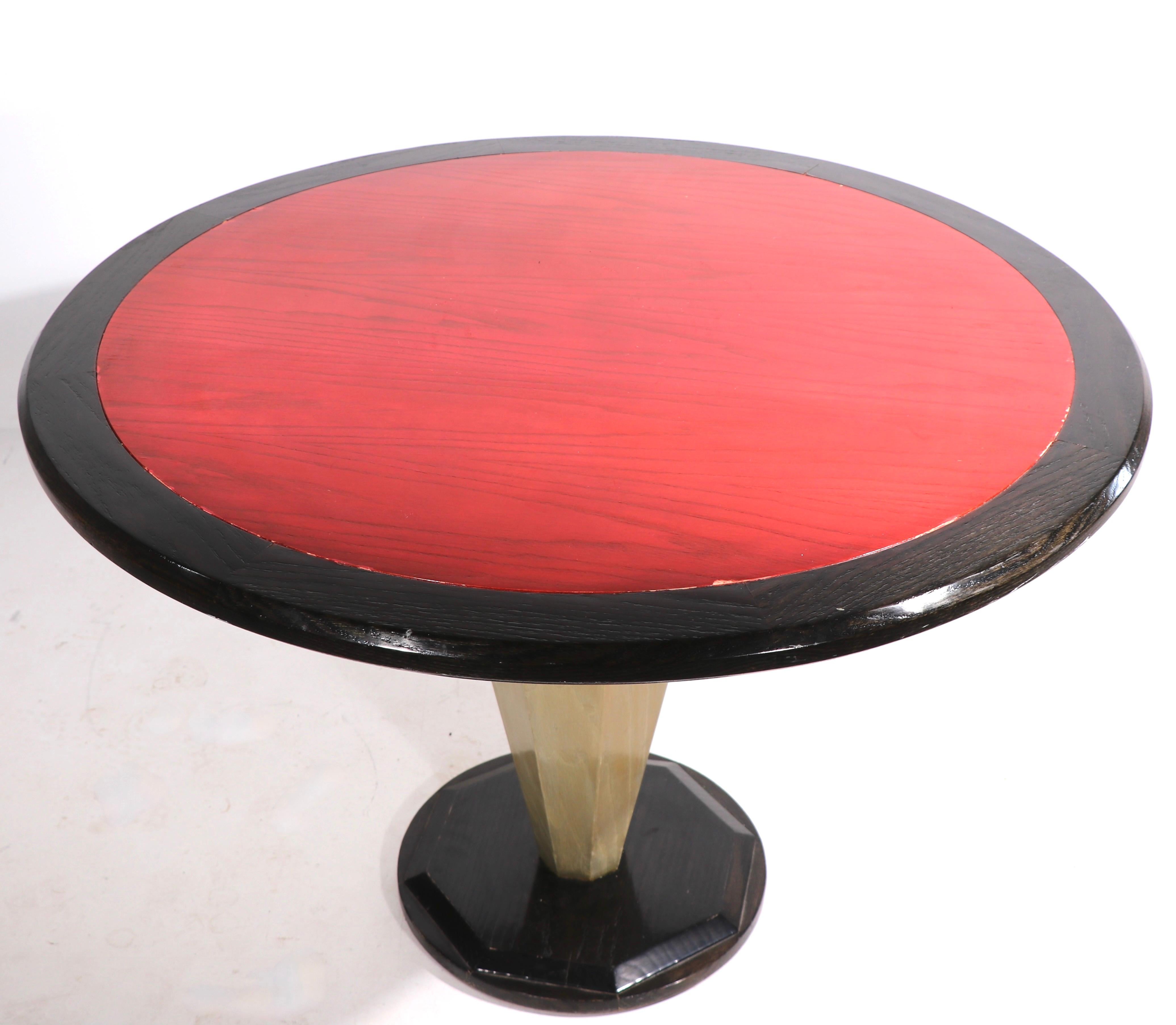Chic round dining, or center, table having an octagonal tapering pedestal center post, which supports the circular top. The top has a red center, with contrasting back trim, the center post os a neutral tan, the stepped base is black. The table