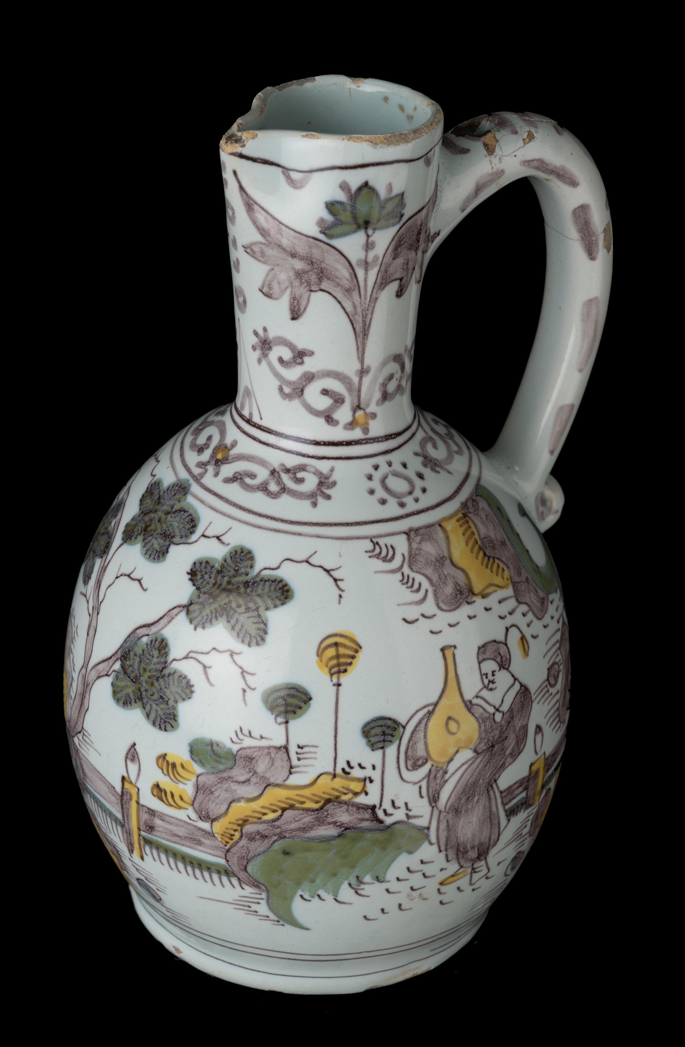 The polychrome wine jug has an ovoid body on a low spreading foot, a conical neck with spout and a handle with a scroll finish. The jug is painted in purple, yellow and green with a continuous chinoiserie landscape with three Chinese figures. A band