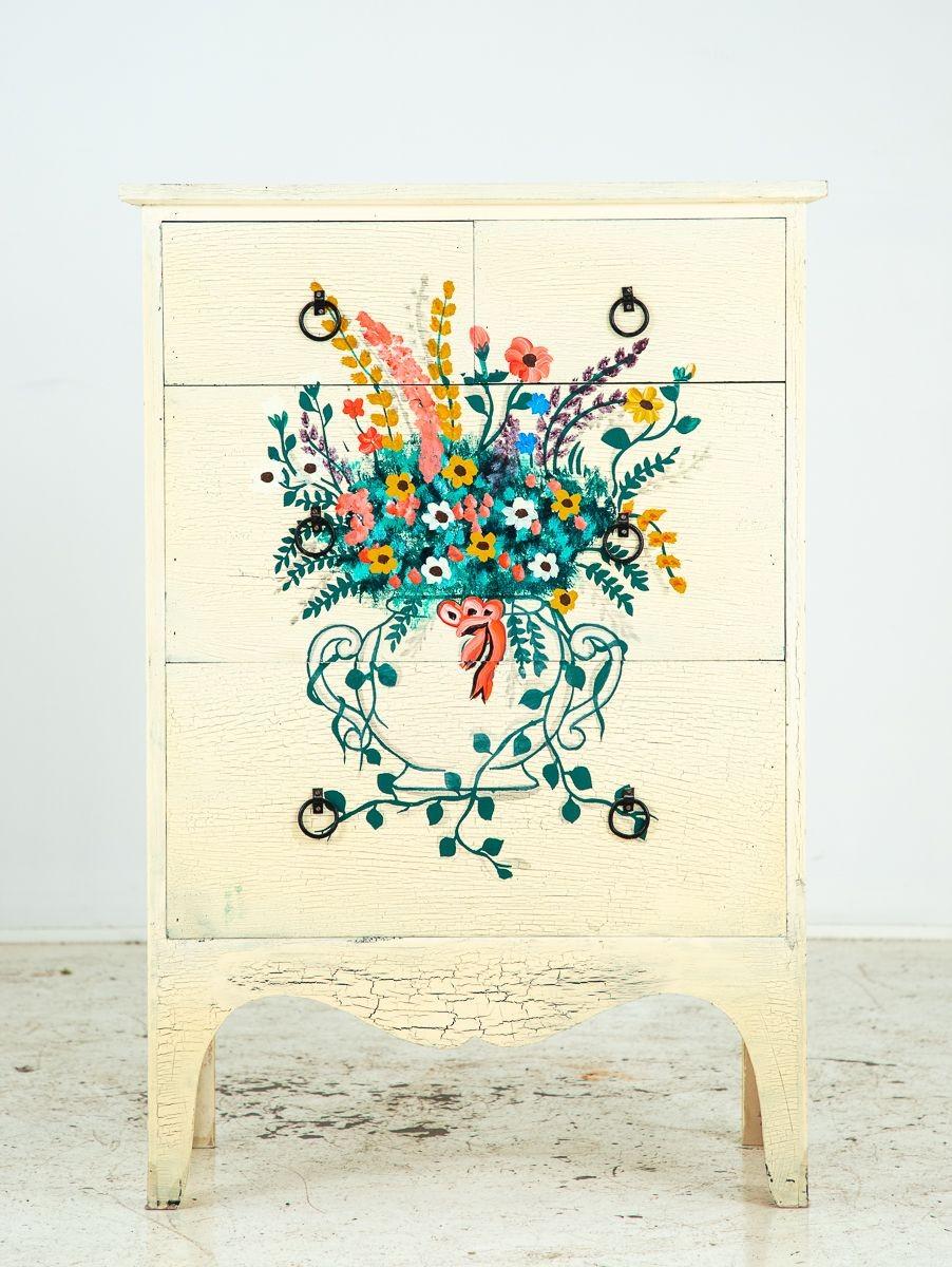 Diese skurrile Kommode zeigt eine handbemalte Vase und ein Blumenarrangement mit kaskadenförmigen Ranken. Der Hintergrund ist in einem warmen Cremegelb gehalten, das die leuchtenden Grün-, Blau-, Rosa-, Violett- und Goldrutentöne hervorhebt. Die