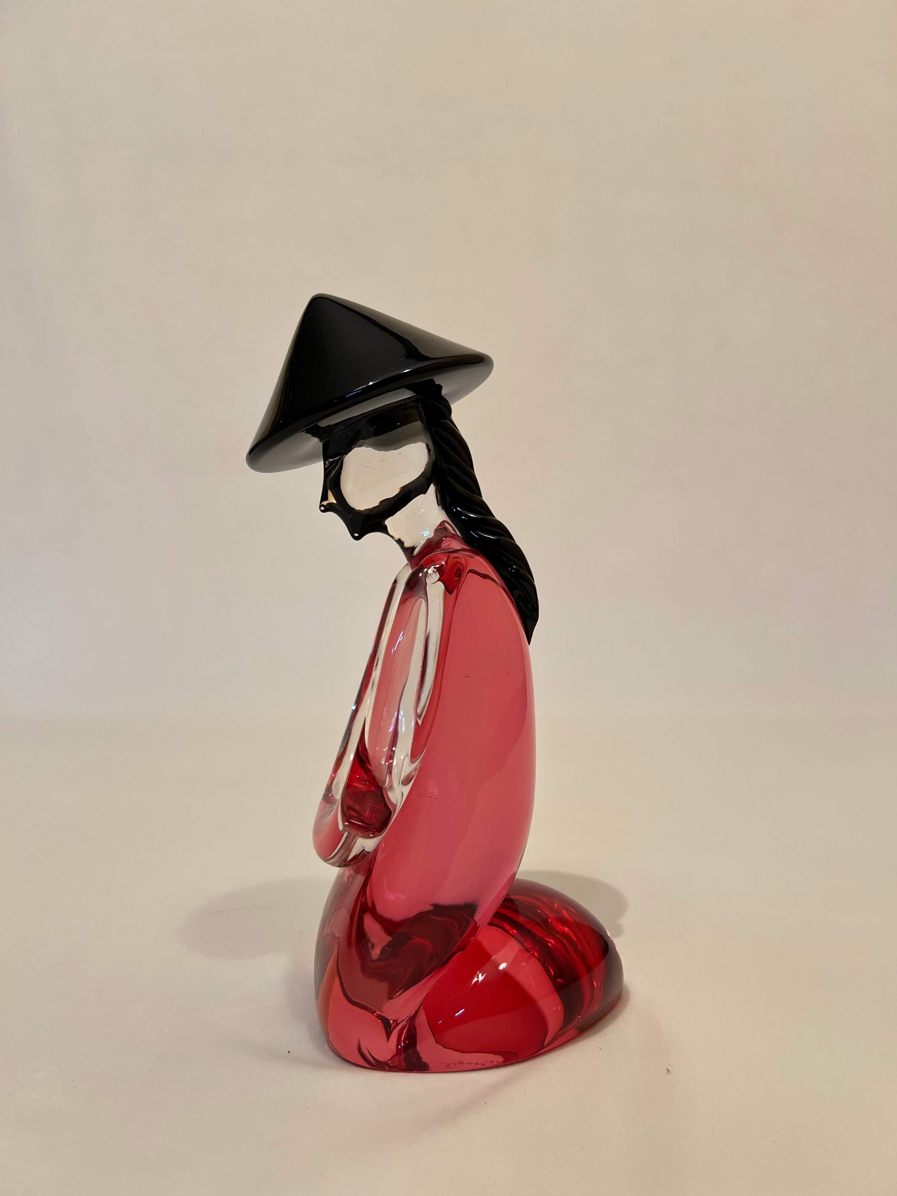 Polychrome statue depicting a kneeling Japanese woman. The statue is in two colours, carmine red and black. The body is red and the typical Japanese hat, and braid, black. The braid has been finely crafted by weaving Murano glass, using ancient