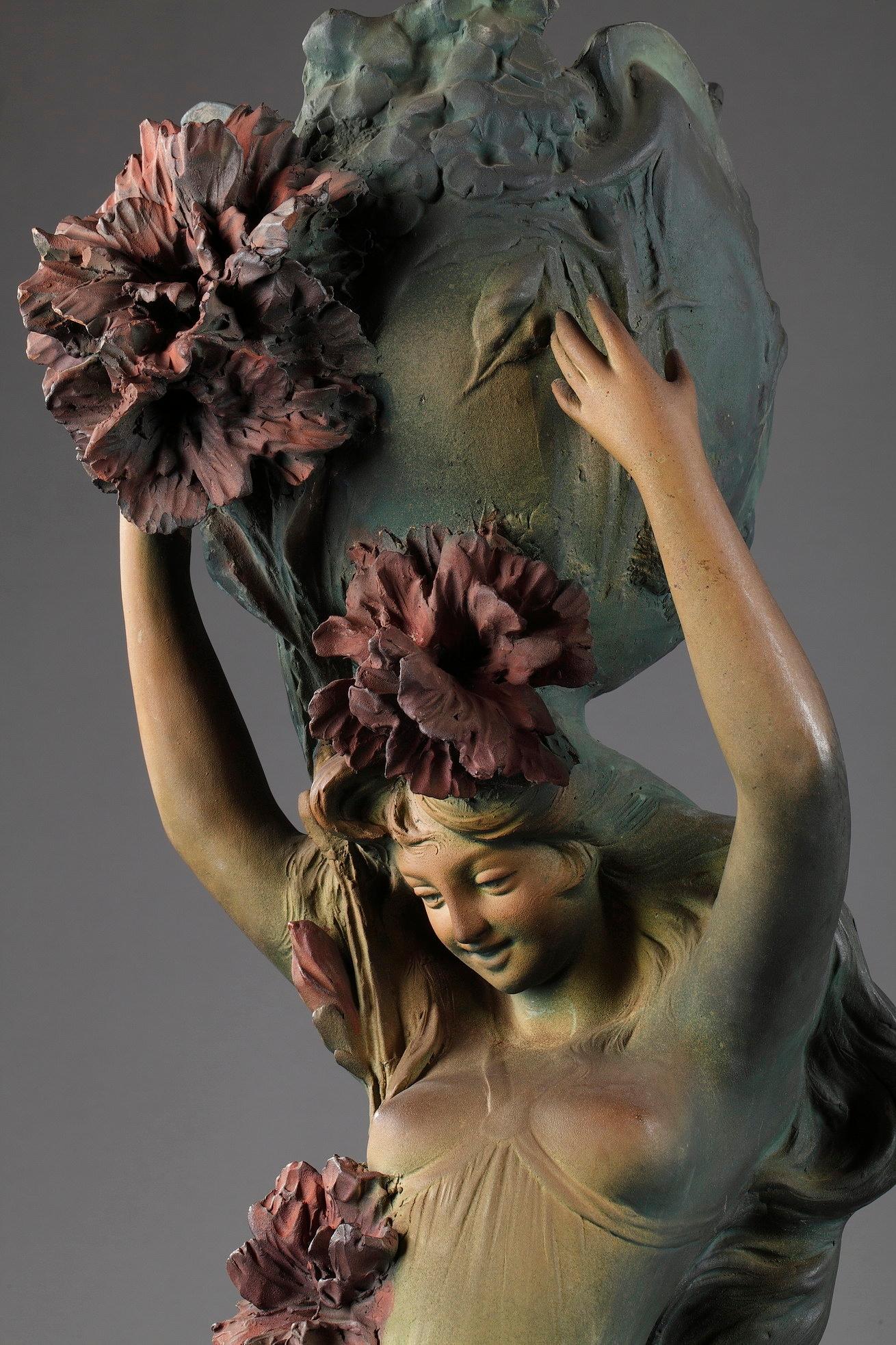 Large polychrome terracotta sculpture and planter. It features a smiling woman wearing a fluid dress that discovers her breasts, holding a basket highlighted with beautiful red flowers. A flowery branch runs along the body of the young woman. The