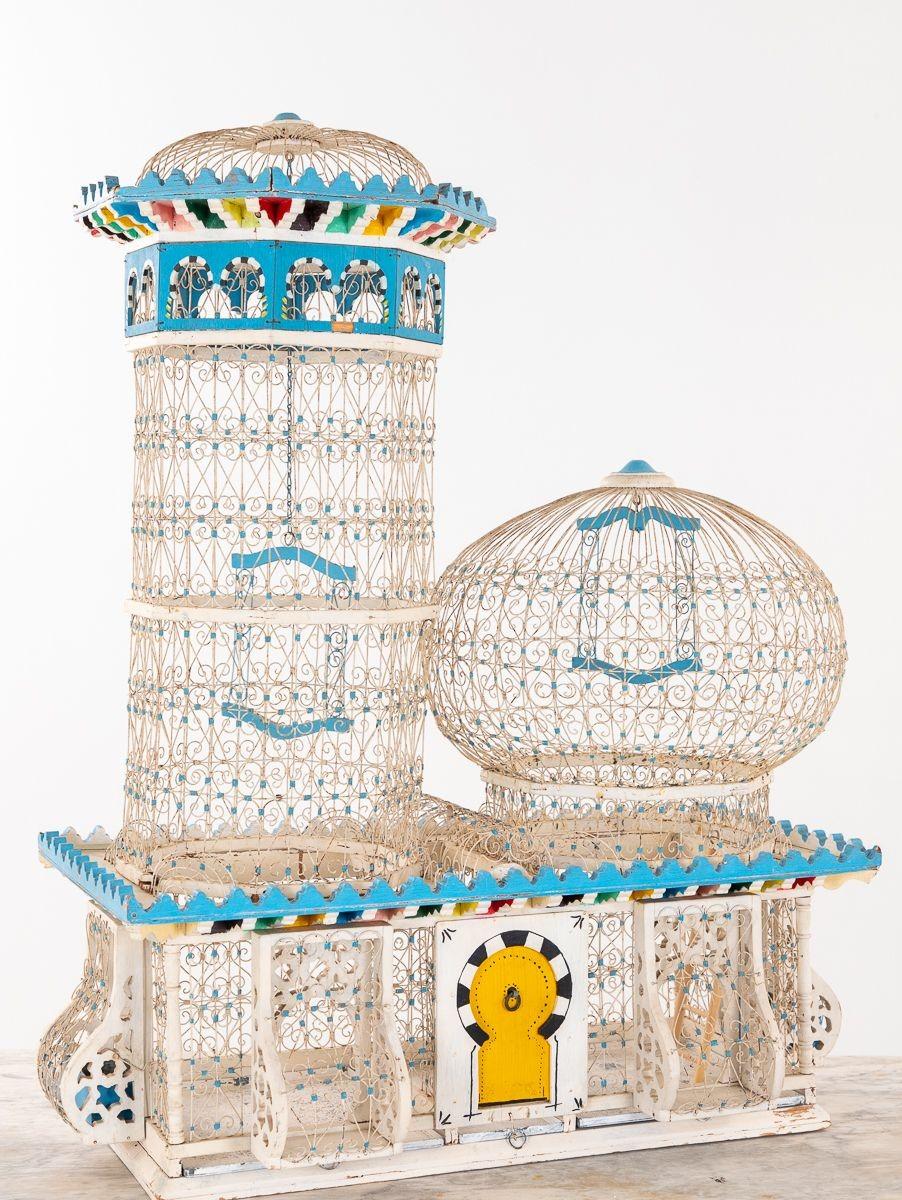 An ornate vintage polychrome wire birdcage featuring a yellow door and two towers. Wooden molding painted in brilliant colors surrounds the crest of the lower level and the taller tower. Each tower features one swing. The wire is shaped to mimic a